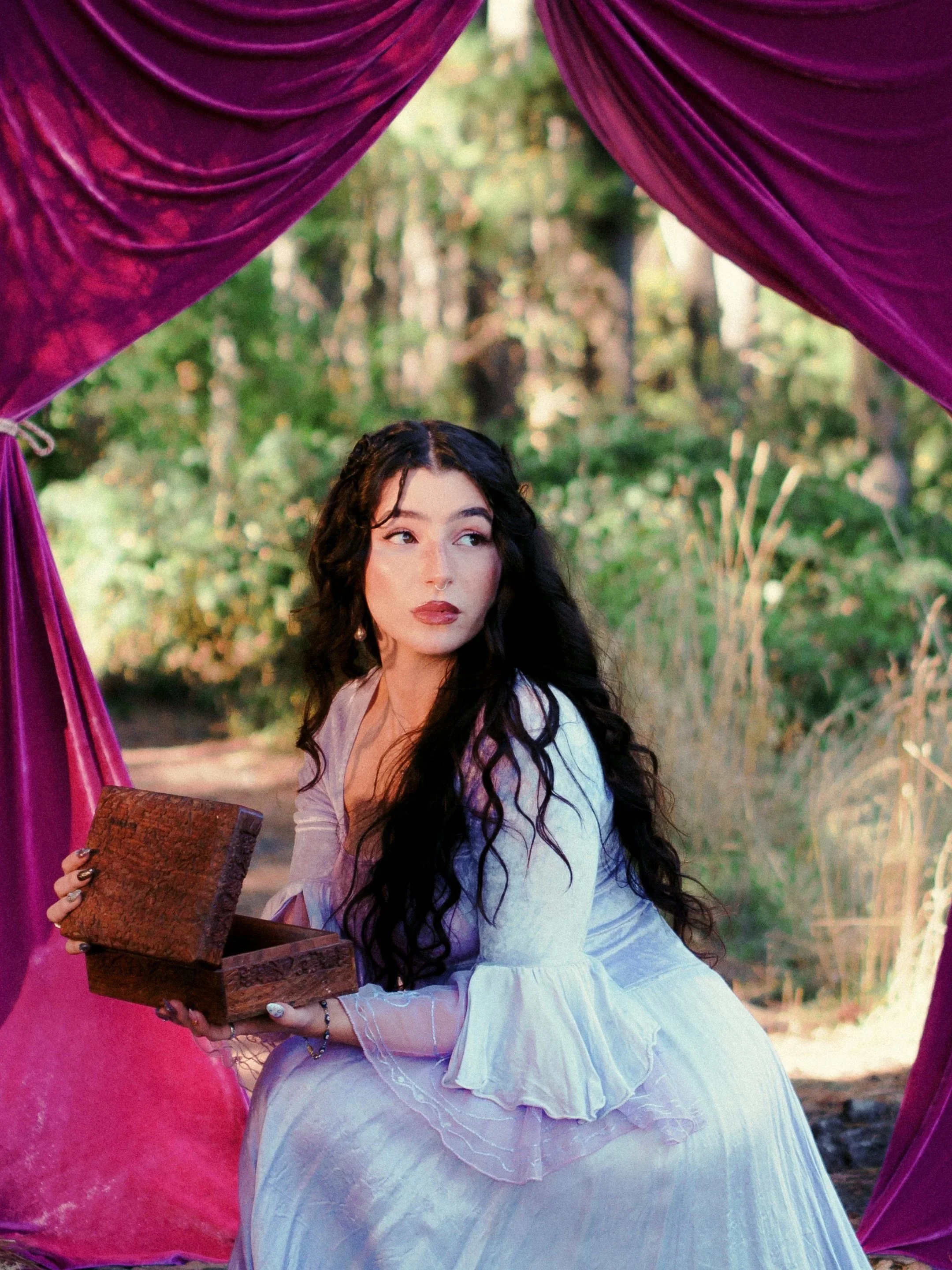 A woman, effie isobel, with dark, curly hair and wearing a white dress, sitting outdoors, viewing into a wooden box. The background shows green trees and sunlight.