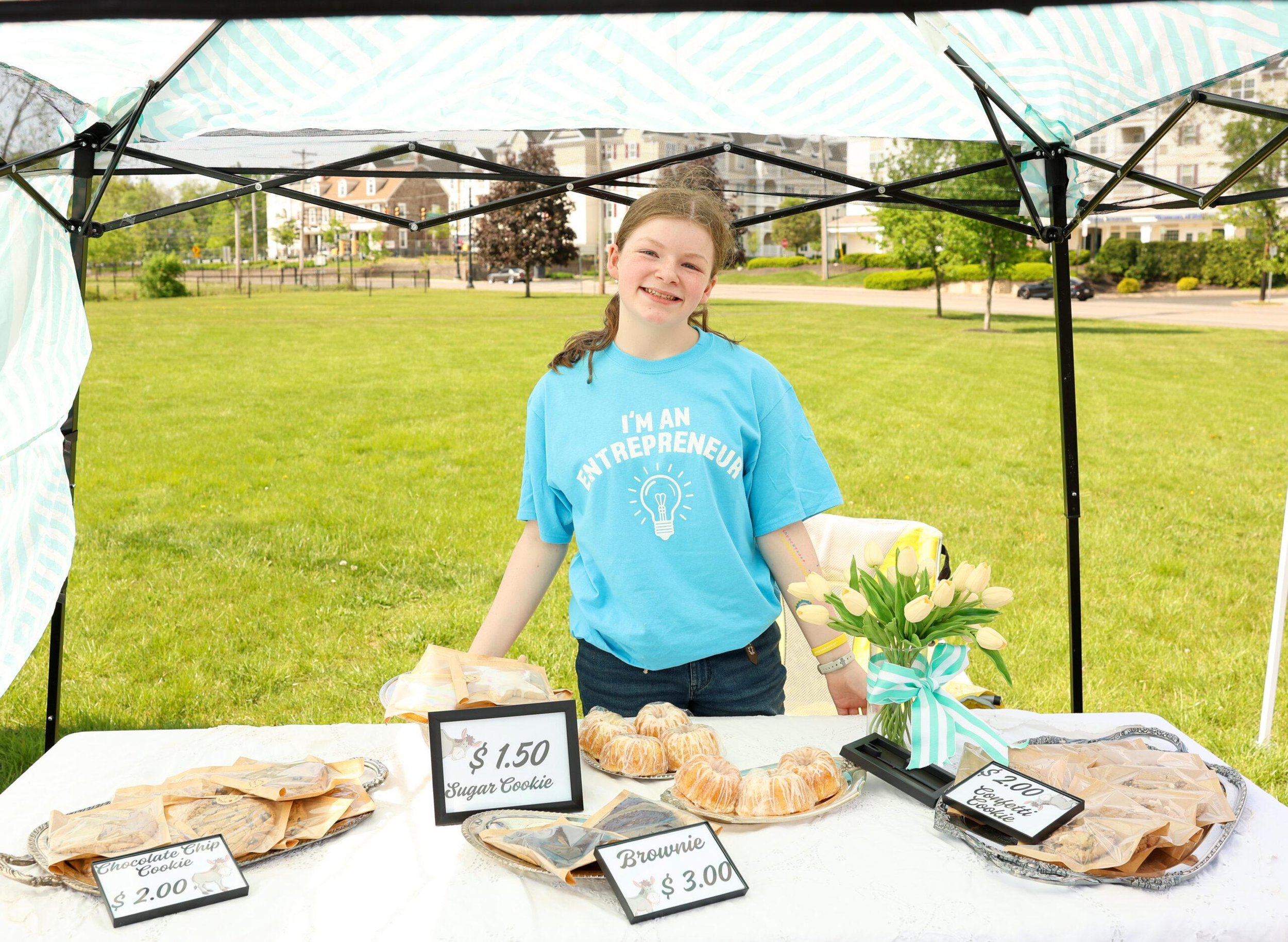 Young girl smiling at a bakery stand selling cookies, with signs showing prices and flavors like chocolate chip, brownie, and sugar cookie, outdoors in a park.