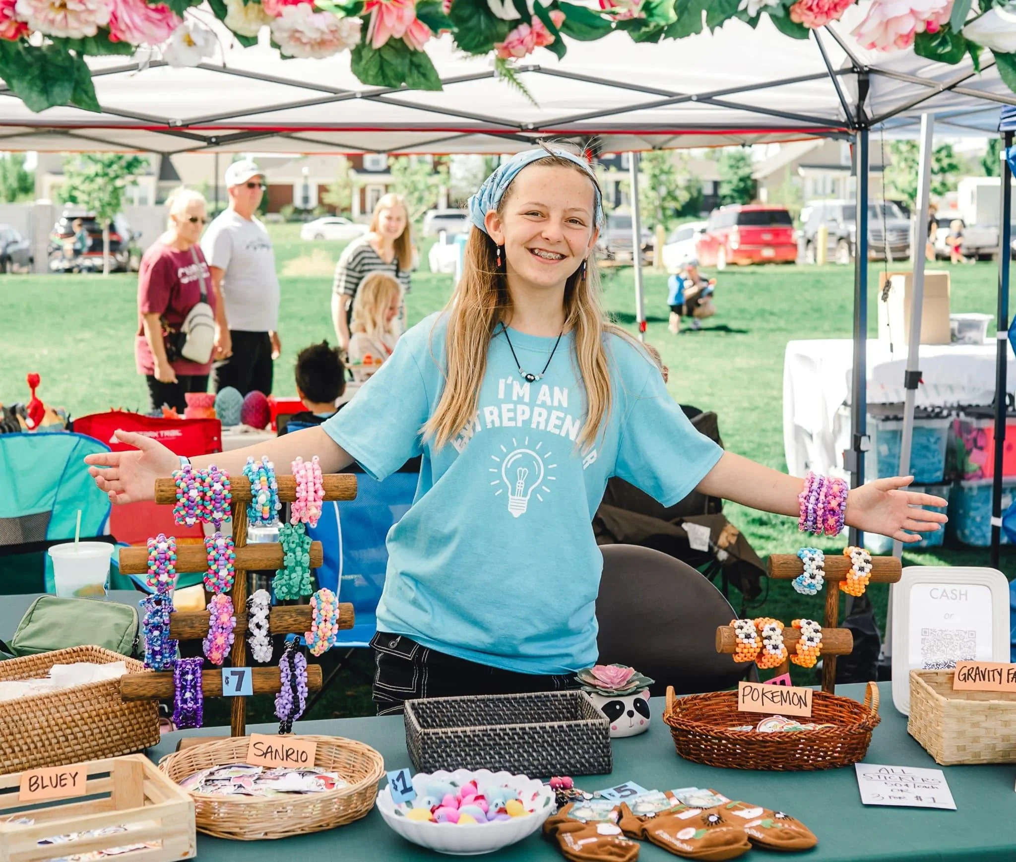 Young girl with long hair and a headband at an outdoor craft fair, smiling with her arms outstretched; stall behind her displays handmade jewelry, plush toys, and crafts, with people browsing in the background.