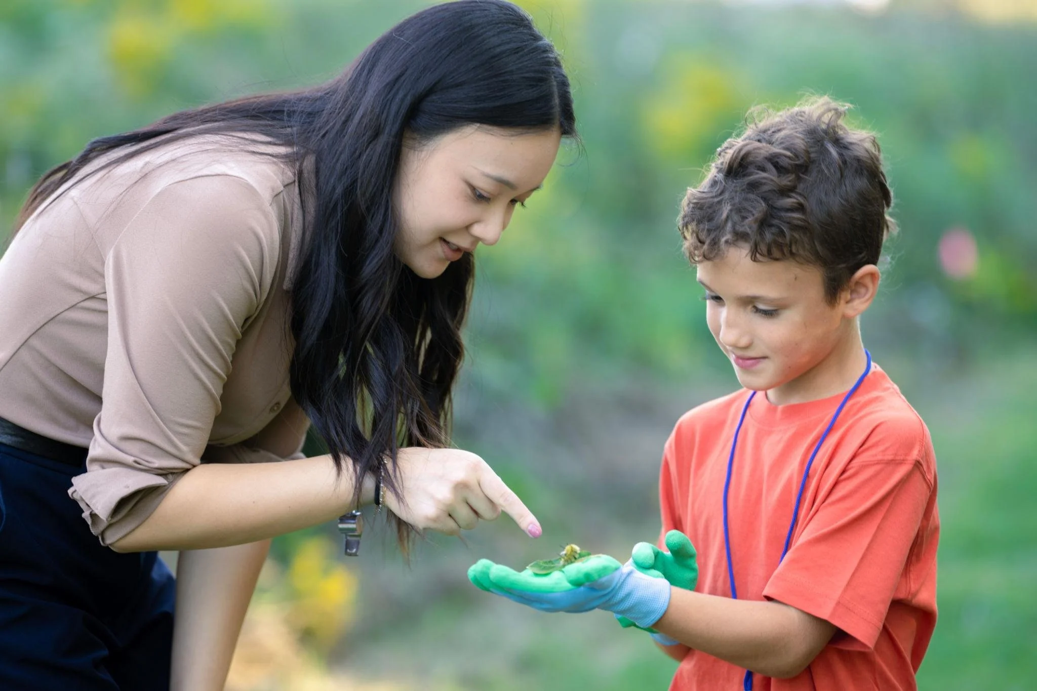 A woman with long dark hair and a young boy with curly hair are outdoors. The woman is pointing at something on the boy’s green gloves, and they appear to be examining an object together on the boy's palm. The background is blurred greenery.