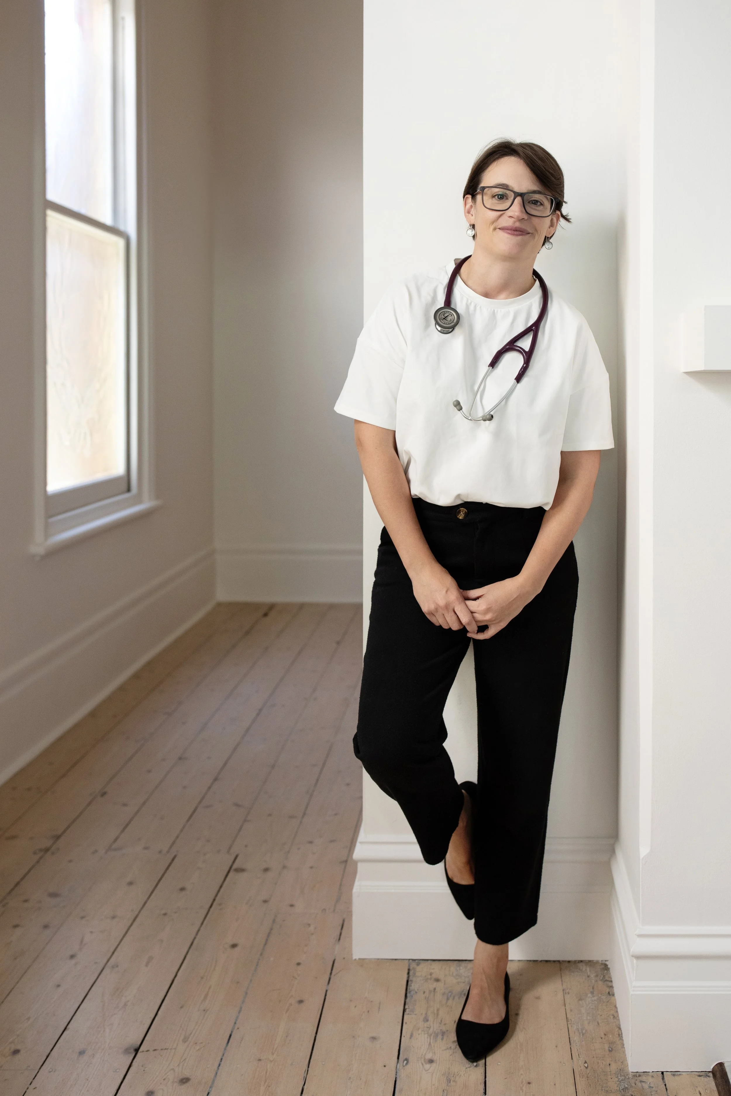A woman with short brown hair, glasses, and earrings wearing a white medical coat and stethoscope around her neck, standing against a white wall in a room with natural light coming from two windows, with her arms crossed and a smile.