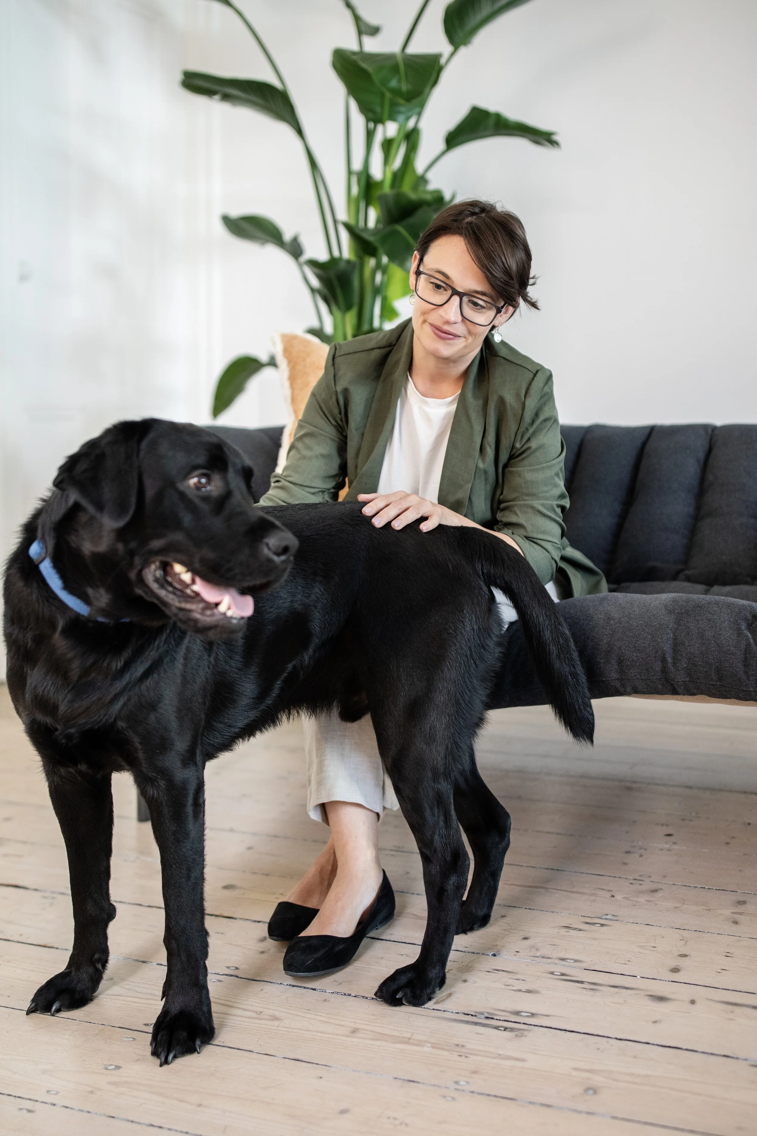 A woman with short brown hair and glasses sitting on a couch, gently petting a large black dog with a blue collar. The dog has its mouth open and tongue slightly out. There is a large green plant behind them and the floor is light-colored wood.
