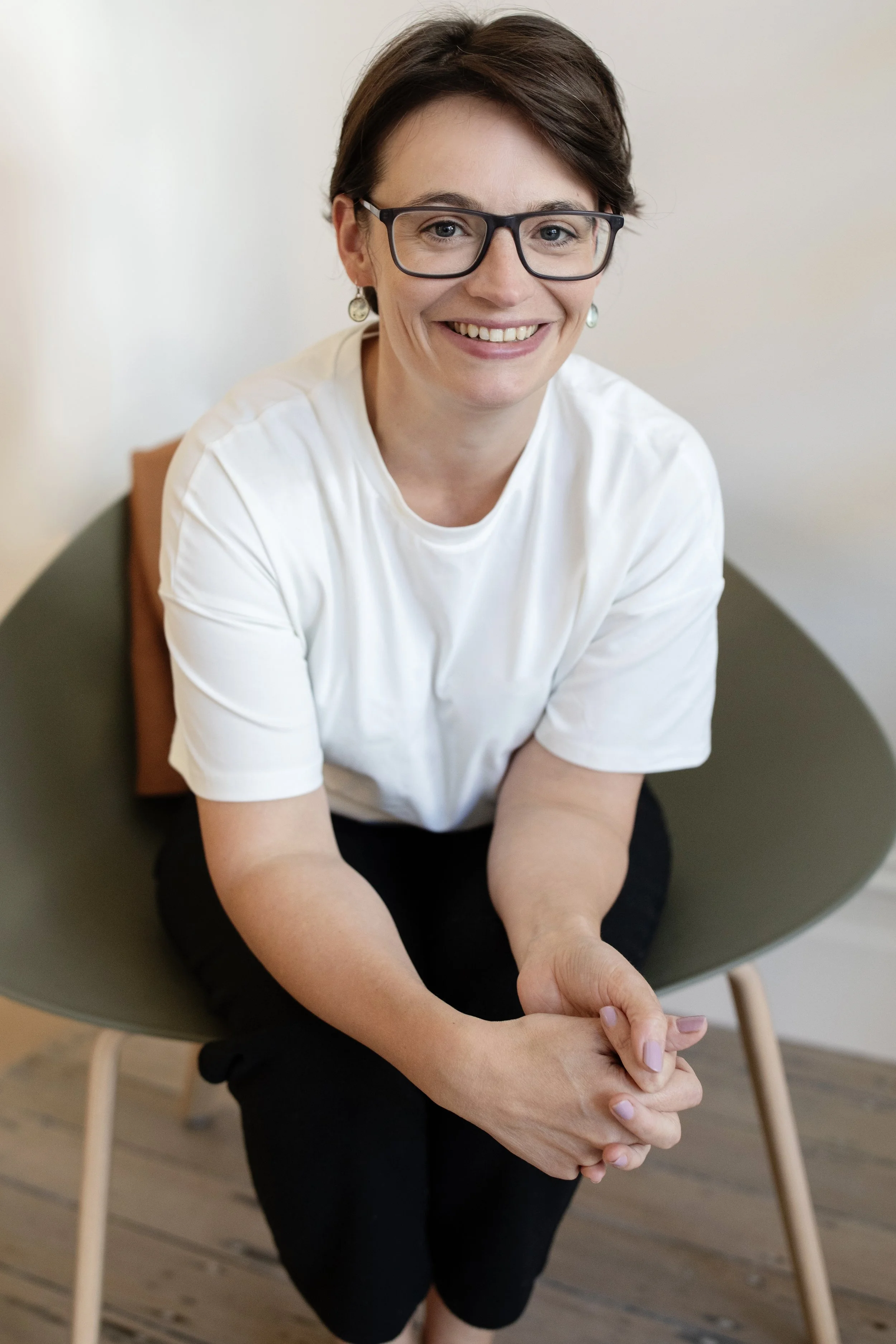 A woman with short brown hair, glasses, and earrings, smiling, sitting on a chair with her arms resting and her hands clasped.