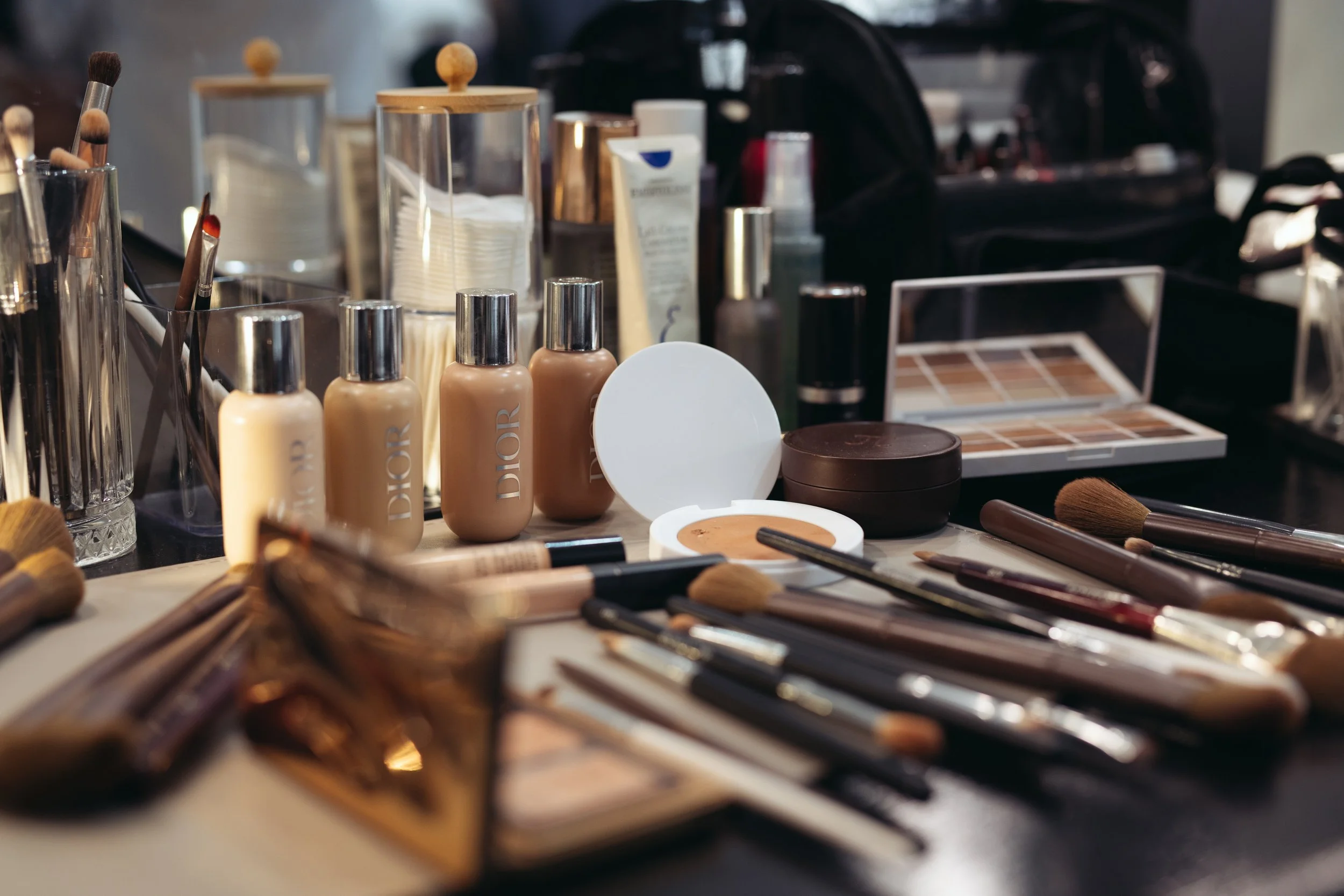 A makeup and beauty station with various cosmetics, brushes, foundation bottles, eyeshadow palettes, and other beauty tools arranged on a table.