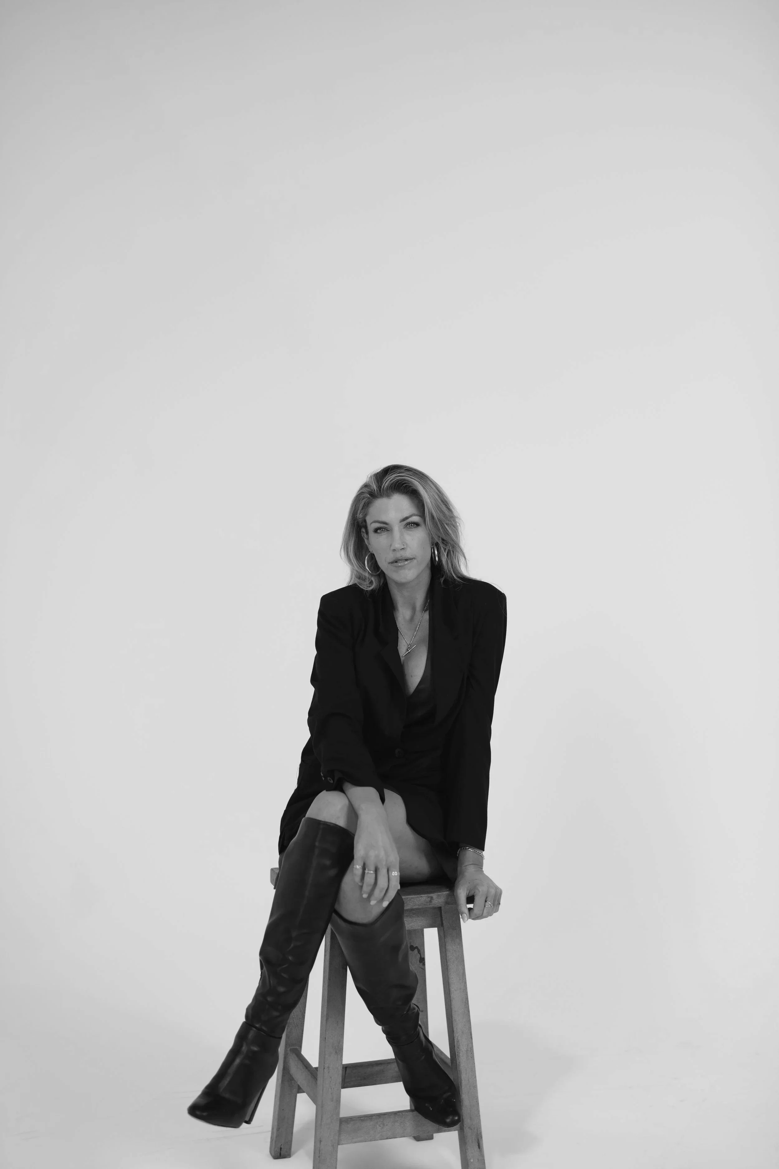 A woman with blonde hair and hoop earrings sitting on a wooden stool against a plain white background. She is wearing a black blazer, black skirt, and thigh-high black leather boots. She looks directly at the camera with a confident expression.