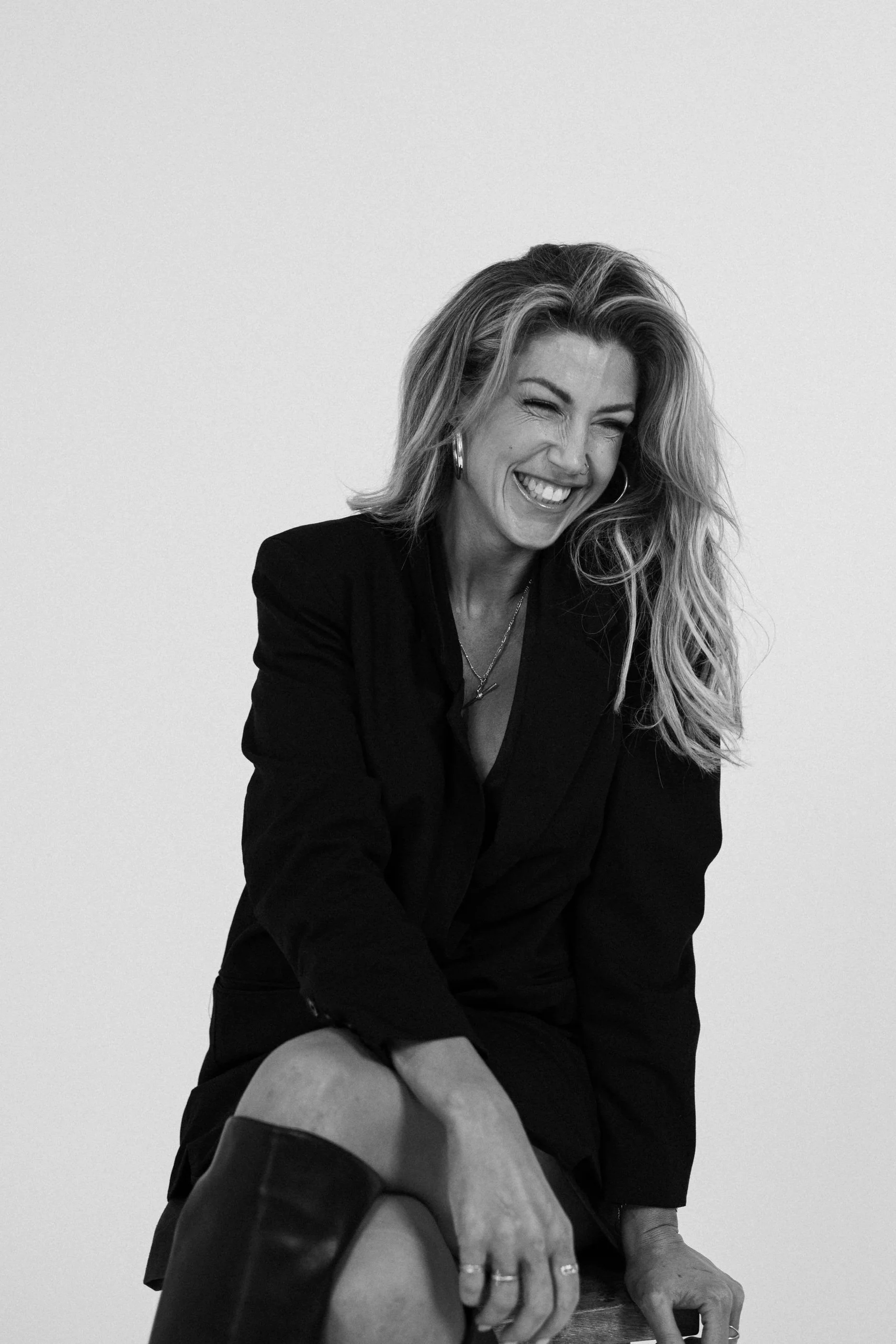 A woman with long wavy hair, smiling and winking, wearing a black blazer, earrings, and rings, sitting on a wooden surface against a plain background.