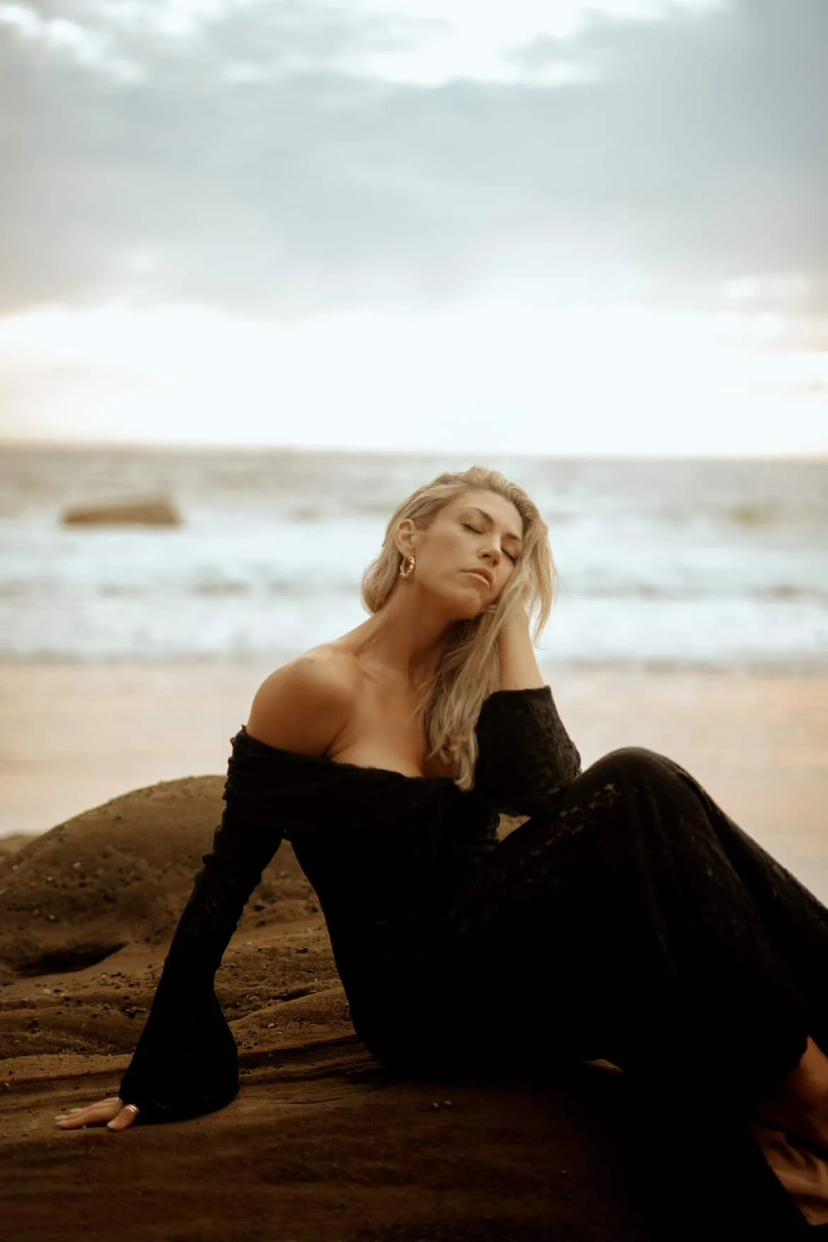 A woman with blonde hair sitting on a rocky beach, wearing a black off-shoulder dress, with her eyes closed, during sunset or sunrise with the ocean in the background.