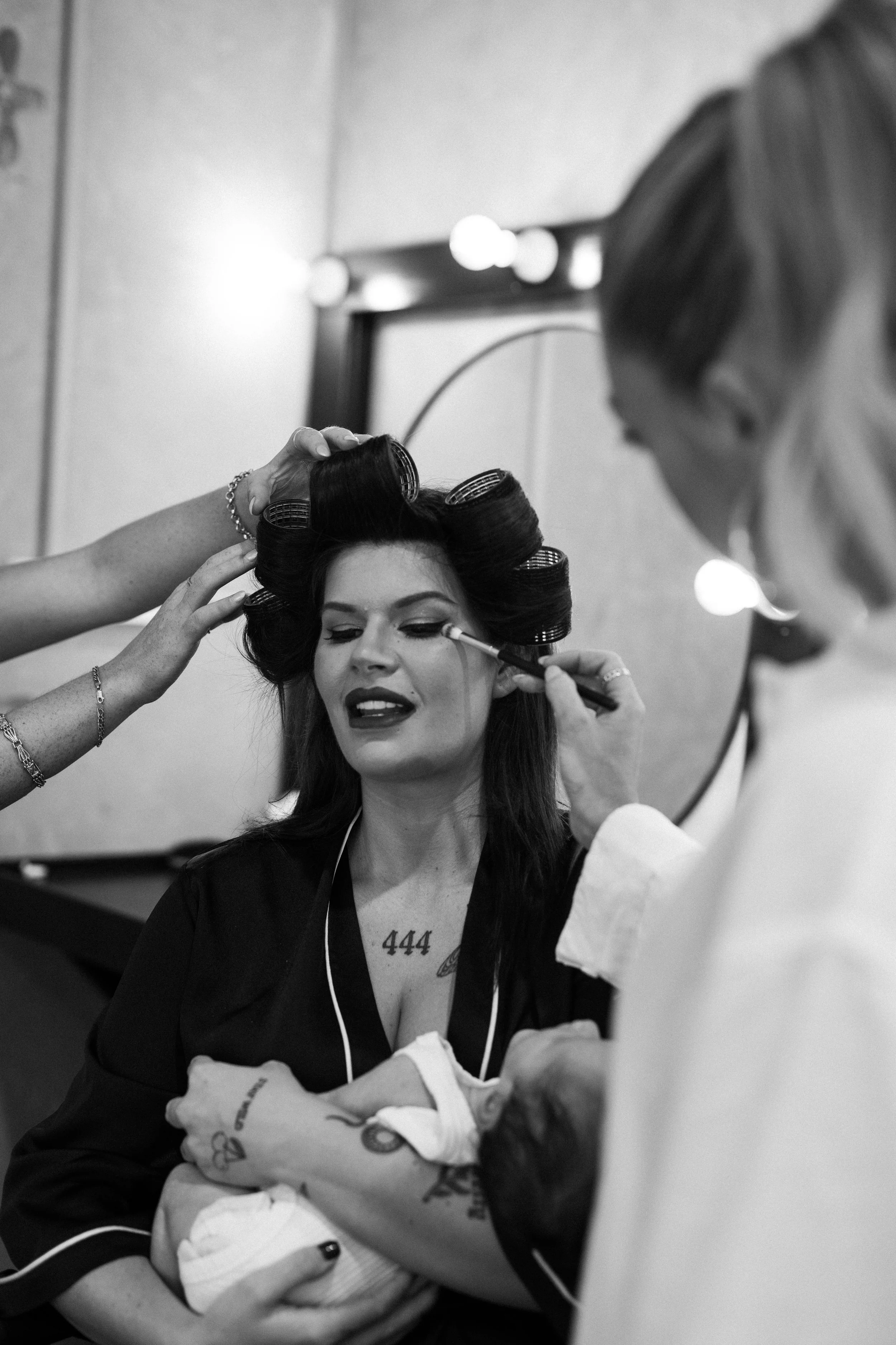 A woman with curlers in her hair and makeup in progress, being helped by a stylist or makeup artist, with a mirror and lights in the background.