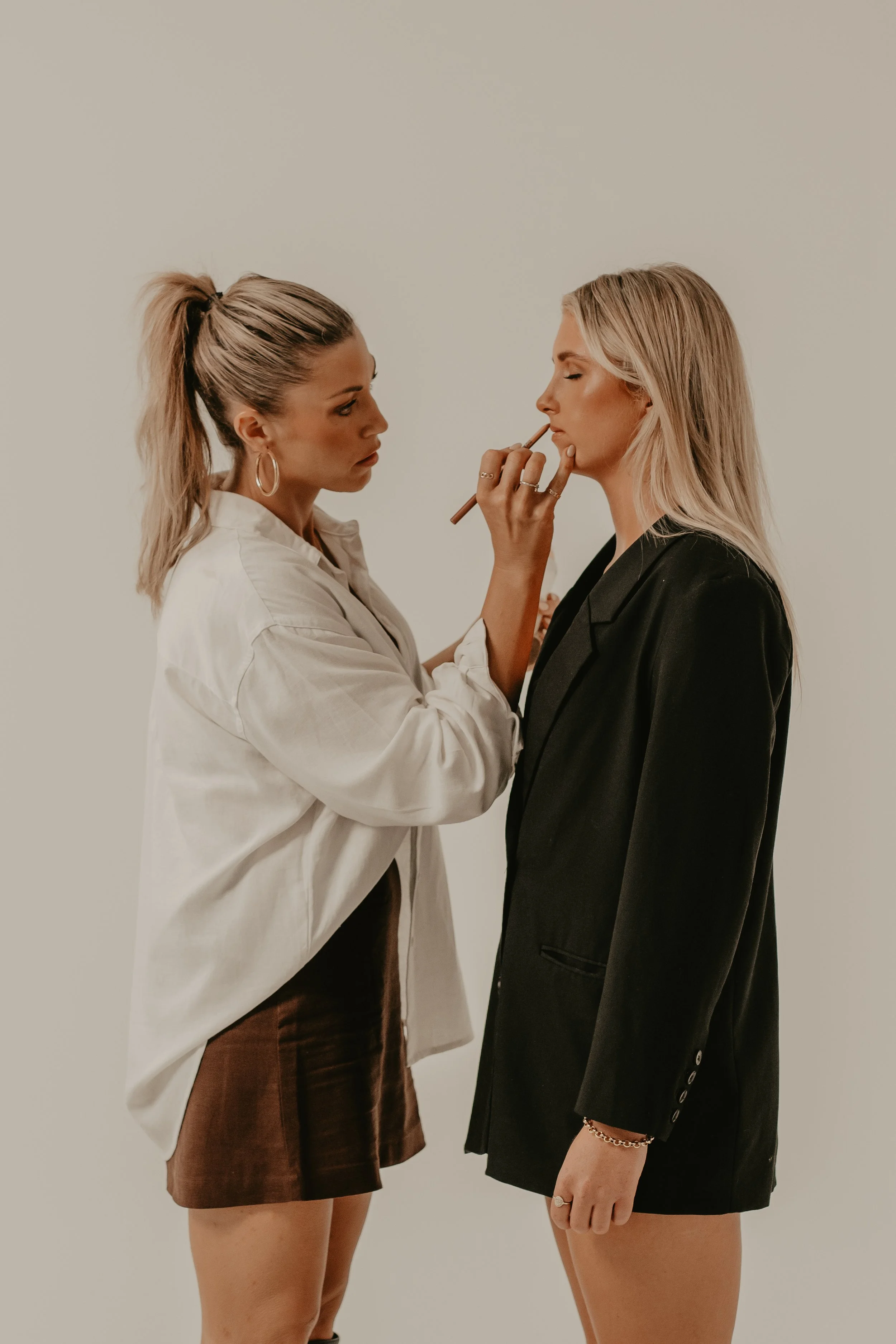 Two women are standing very close to each other, one is applying makeup to the other, against a plain wall background.