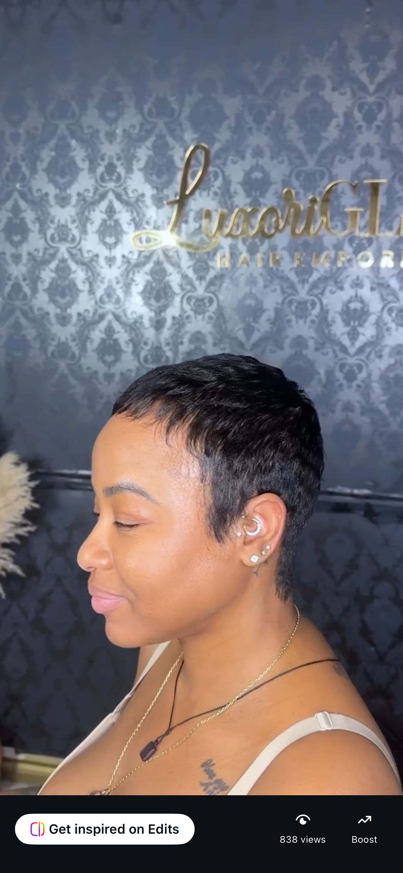 Side profile of a woman with short, textured black hairstyle, multiple earrings, and wearing layered necklaces inside a salon, with a decorative wallpaper and a sign that reads 'Luvace L Hair & Beauty' in the background.