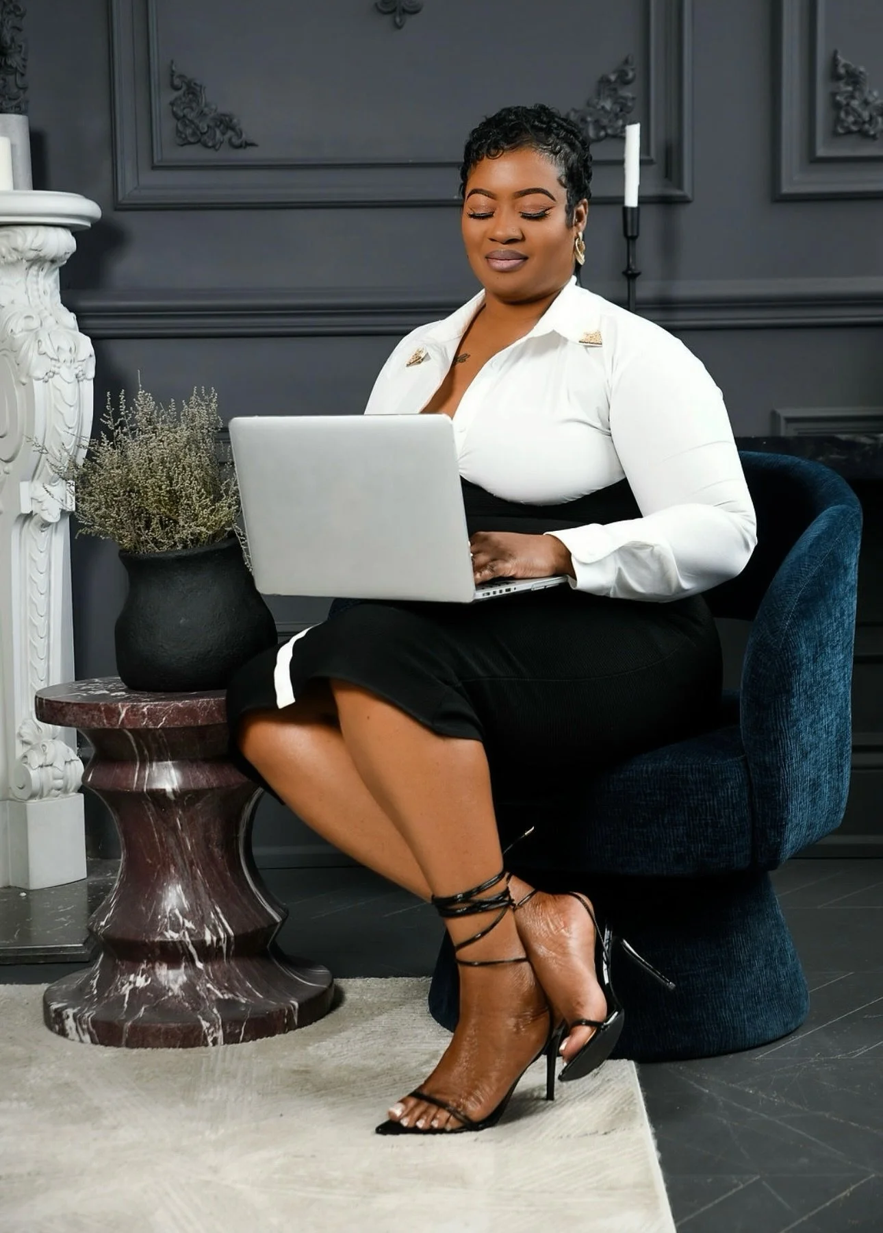 A woman sitting in a dark blue armchair, working on a silver laptop in a stylish room with dark gray walls, decorative moldings, and candle sconces. She wears a black skirt, white blouse with gold accents, and brown strappy high heels, with her legs crossed and eyes closed.