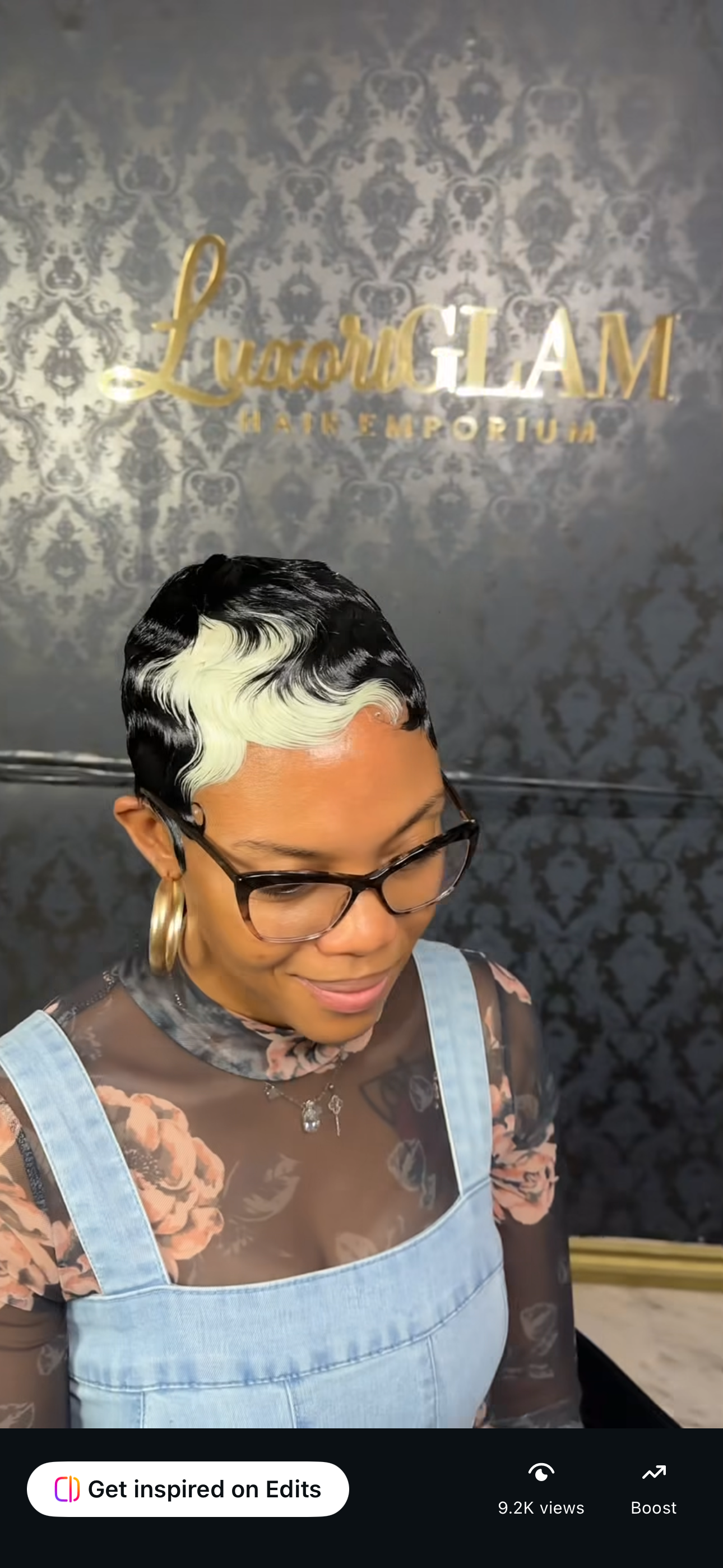 A woman with short black hair styled with blonde waves at the front, wearing glasses, hoop earrings, a floral top, and denim overalls, standing in front of a dark patterned wall with a gold sign reading "Luxury Lavi Hair & Emporium".