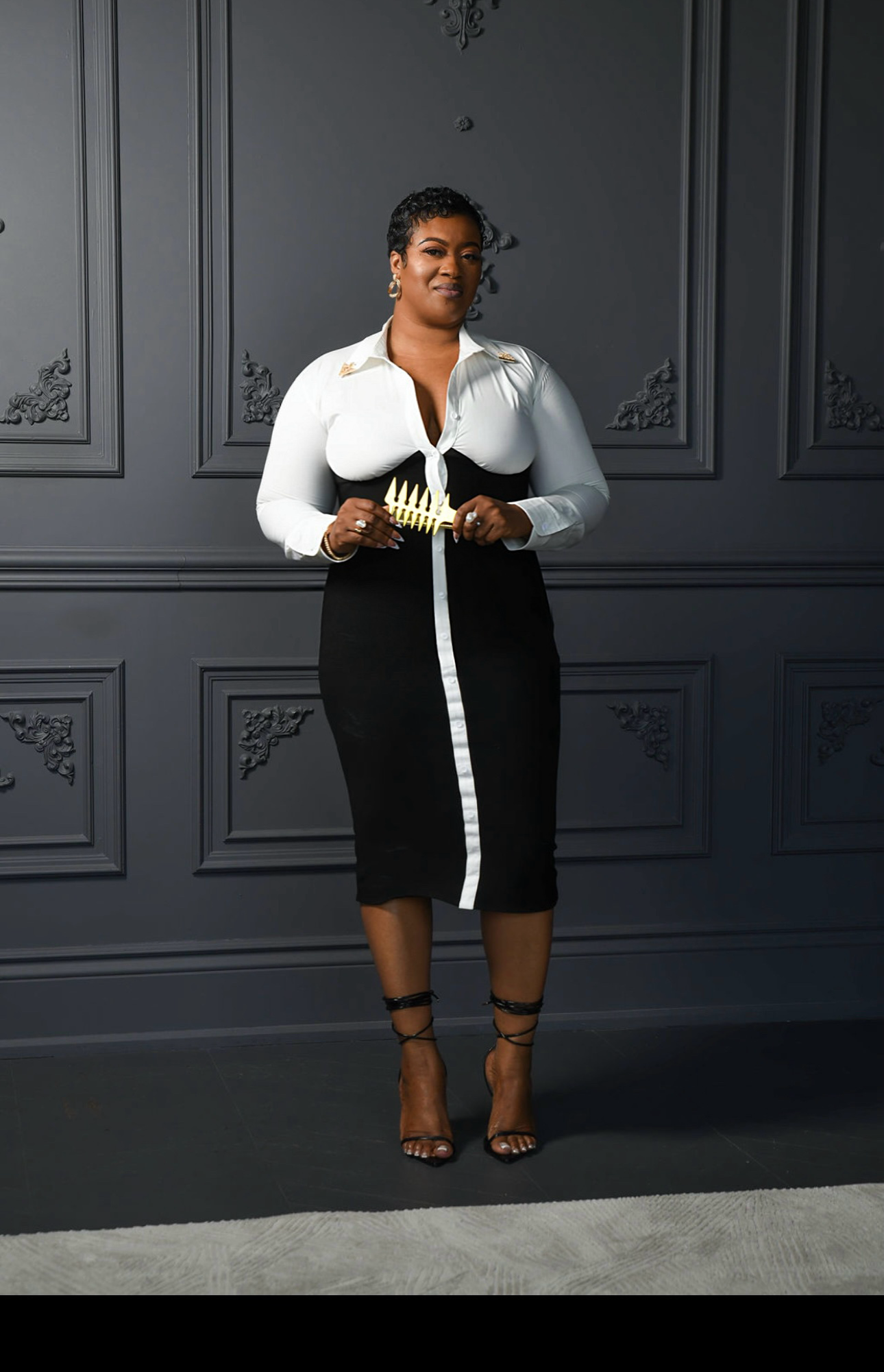 A woman in a black and white outfit standing in front of a dark gray wall with decorative molding, holding a gold trident-shaped award, wearing high heels.