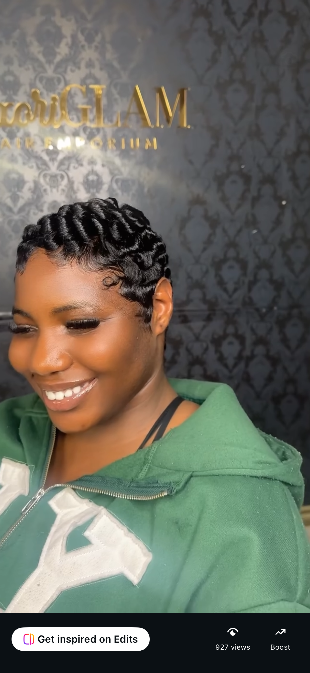 A woman with a short, textured hairstyle, smiling, wearing a green hoodie, and standing in front of a dark patterned wall with gold lettering.