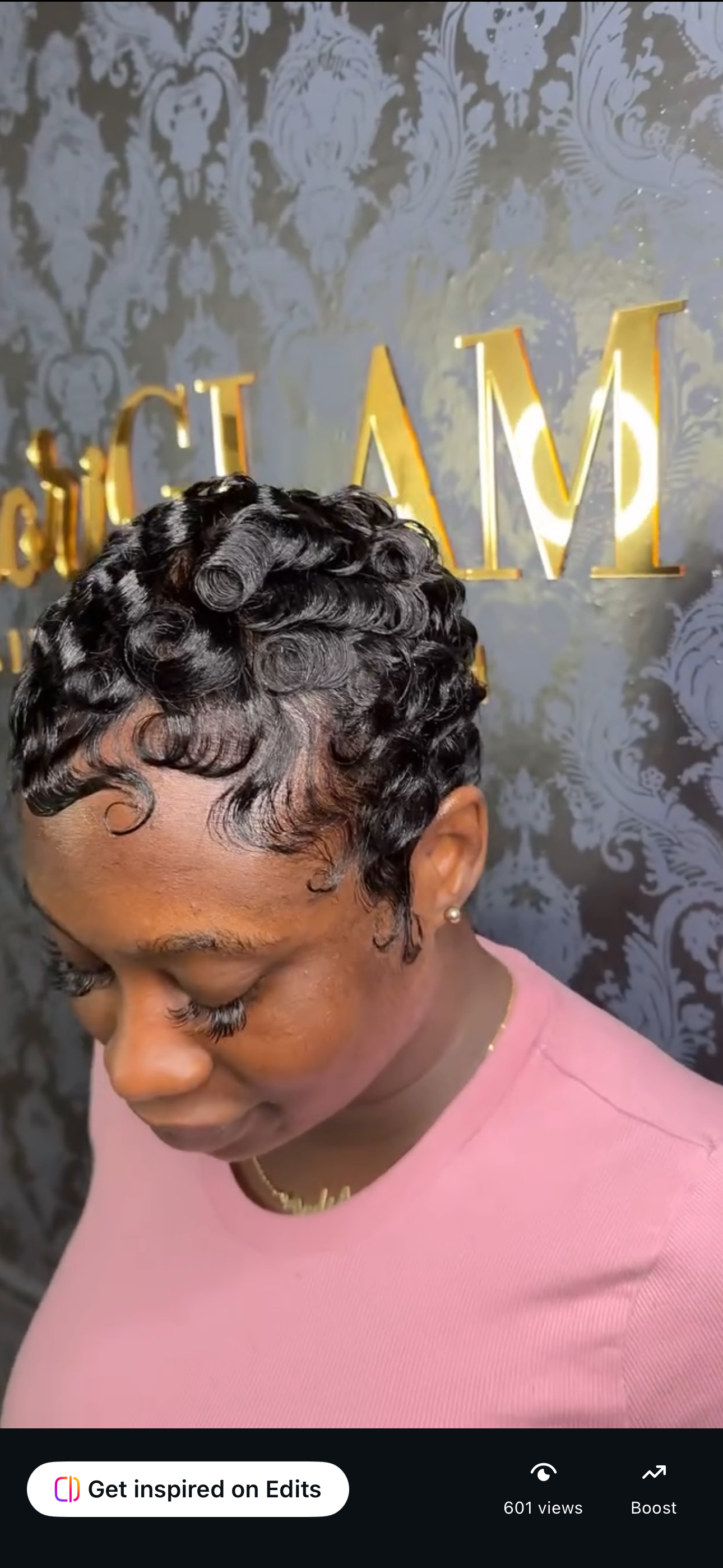 Close-up of a woman with short, curly black hair styled in defined curls, wearing a pink top, with a dark patterned wall and gold letters in the background.