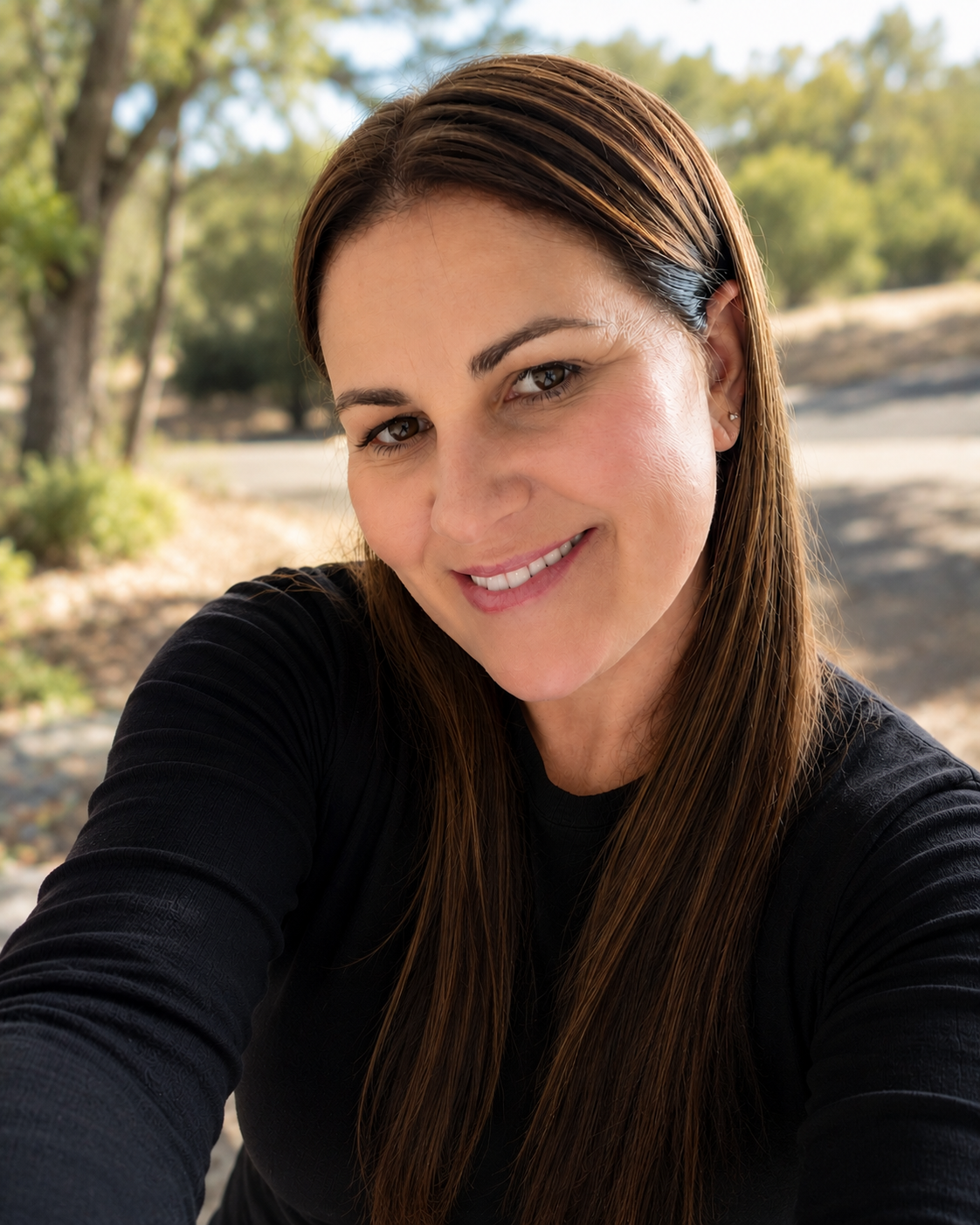 Close-up of Jacinda Stewart, founder of Run Recruitment. Smiling woman with long brown hair, wearing a black top, outdoors in a park with trees and sunlight. Expert in writing professional resumes and an experienced recruiter.