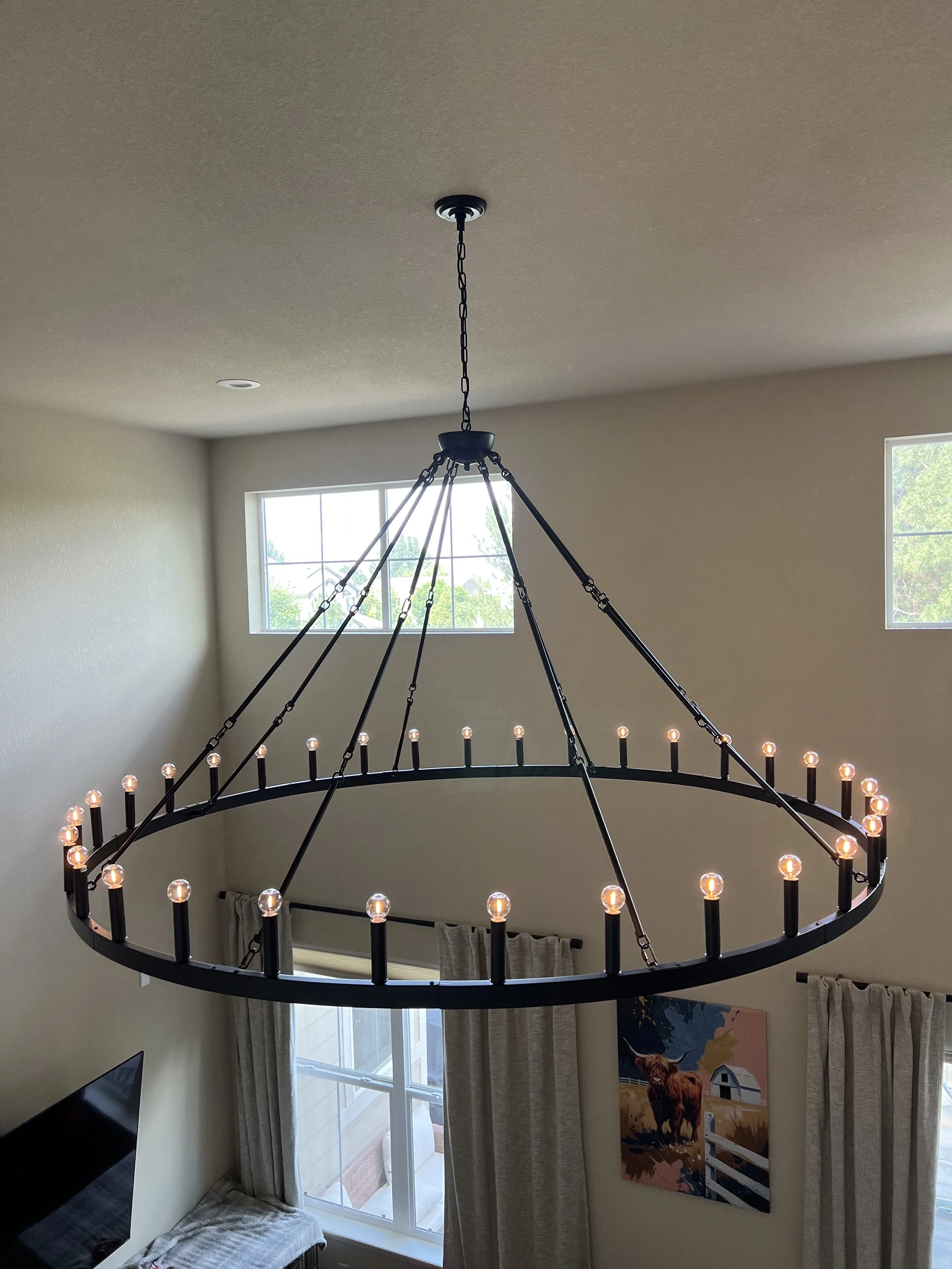 A large circular chandelier with numerous small light bulbs hanging from the ceiling in a living room. The room has two large windows with curtains and a picture of a bull on the wall.