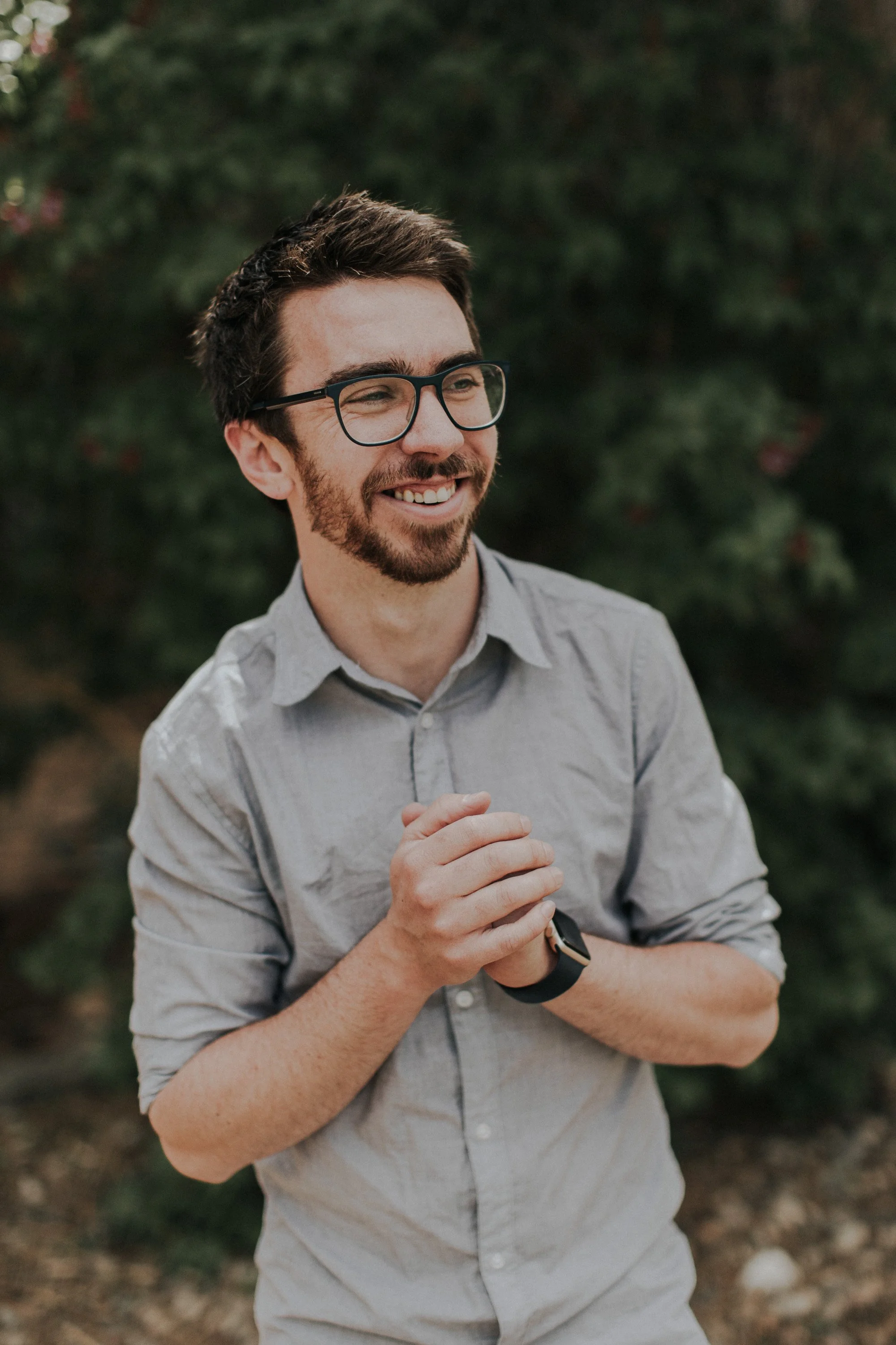 A smiling man with glasses, a beard, and a smartwatch, wearing a light gray shirt, standing outdoors with blurred green foliage in the background.