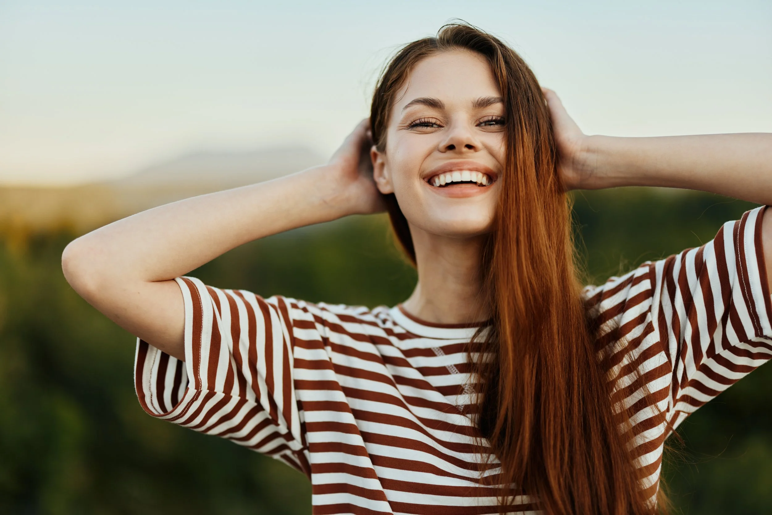 A young woman with long red hair smiling outdoors, wearing a red and white striped shirt, with her hands behind her head against a natural background.