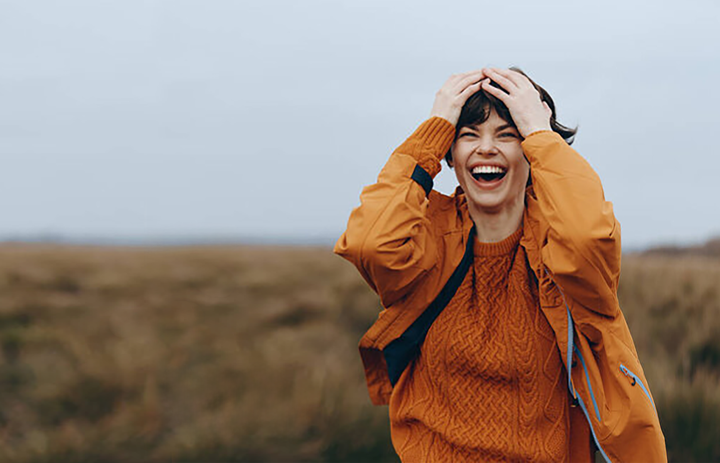 A woman in an orange jacket and sweater is outside on a cloudy day, smiling and laughing with her hands on her head.