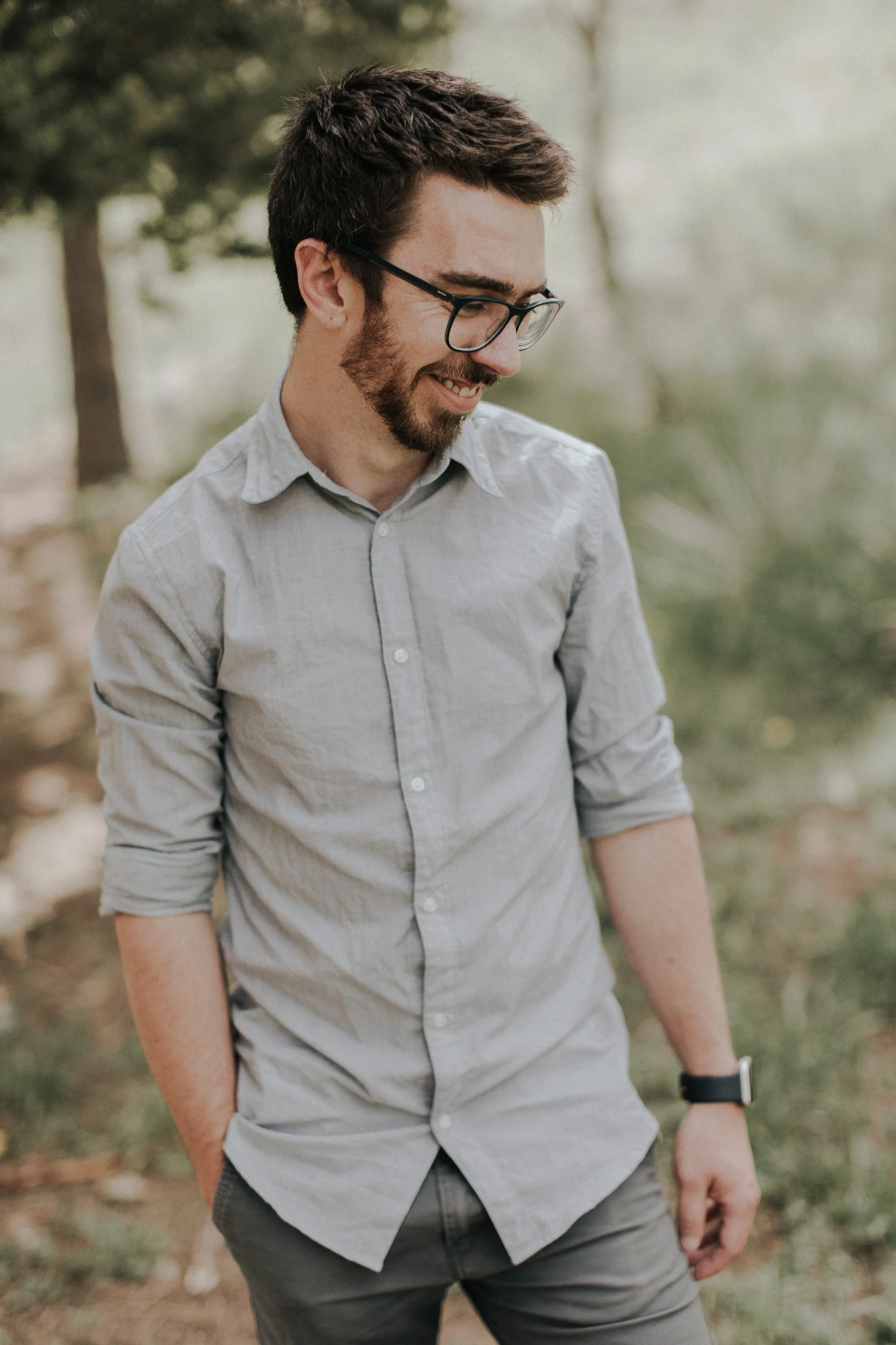 A man with short dark hair, glasses, a beard, and a light gray button-up shirt smiling outdoors, with a blurred natural background.
