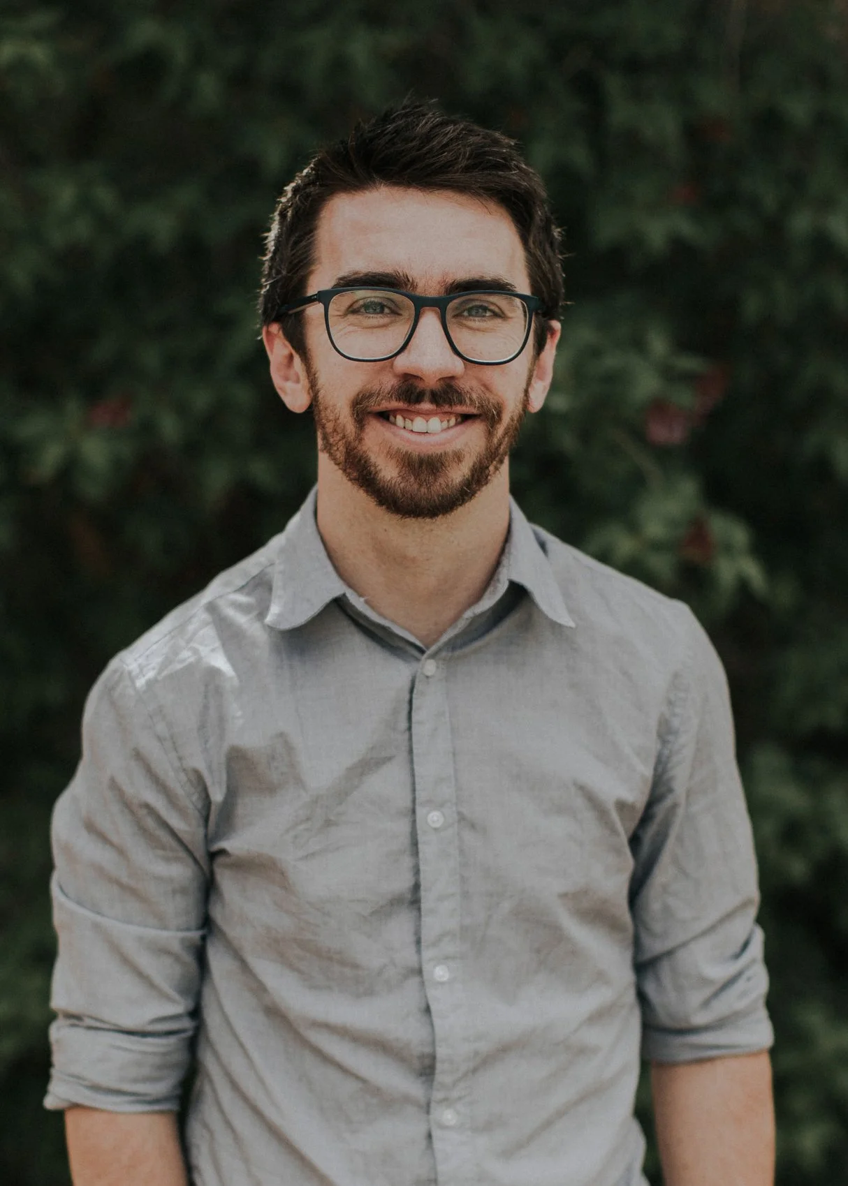 A young man with dark hair, glasses, and a beard, smiling, wearing a gray shirt with rolled-up sleeves, standing outdoors with green foliage in the background.