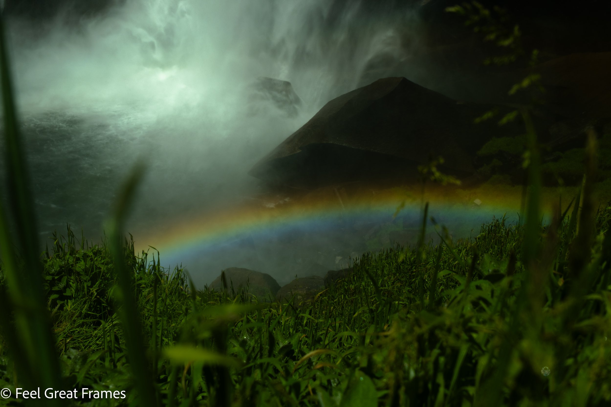 A waterfall cascading down rocks with mist, viewed through green plants and grass, and a rainbow at the base of the waterfall.