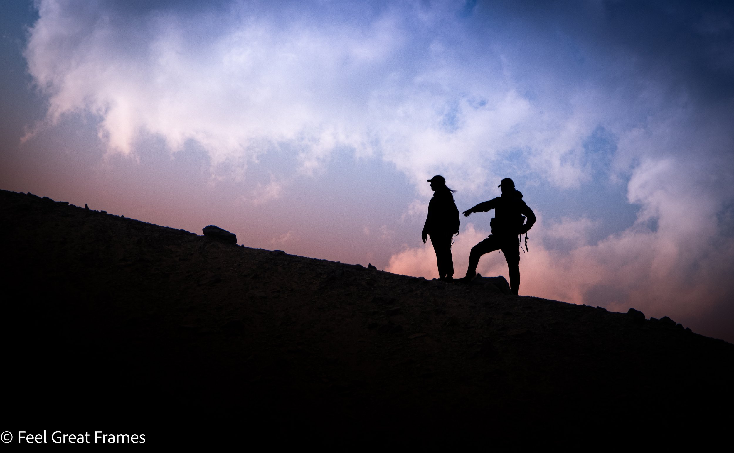 Two silhouetted hikers standing on a mountain ridge during sunset, with a colorful sky filled with clouds in the background.