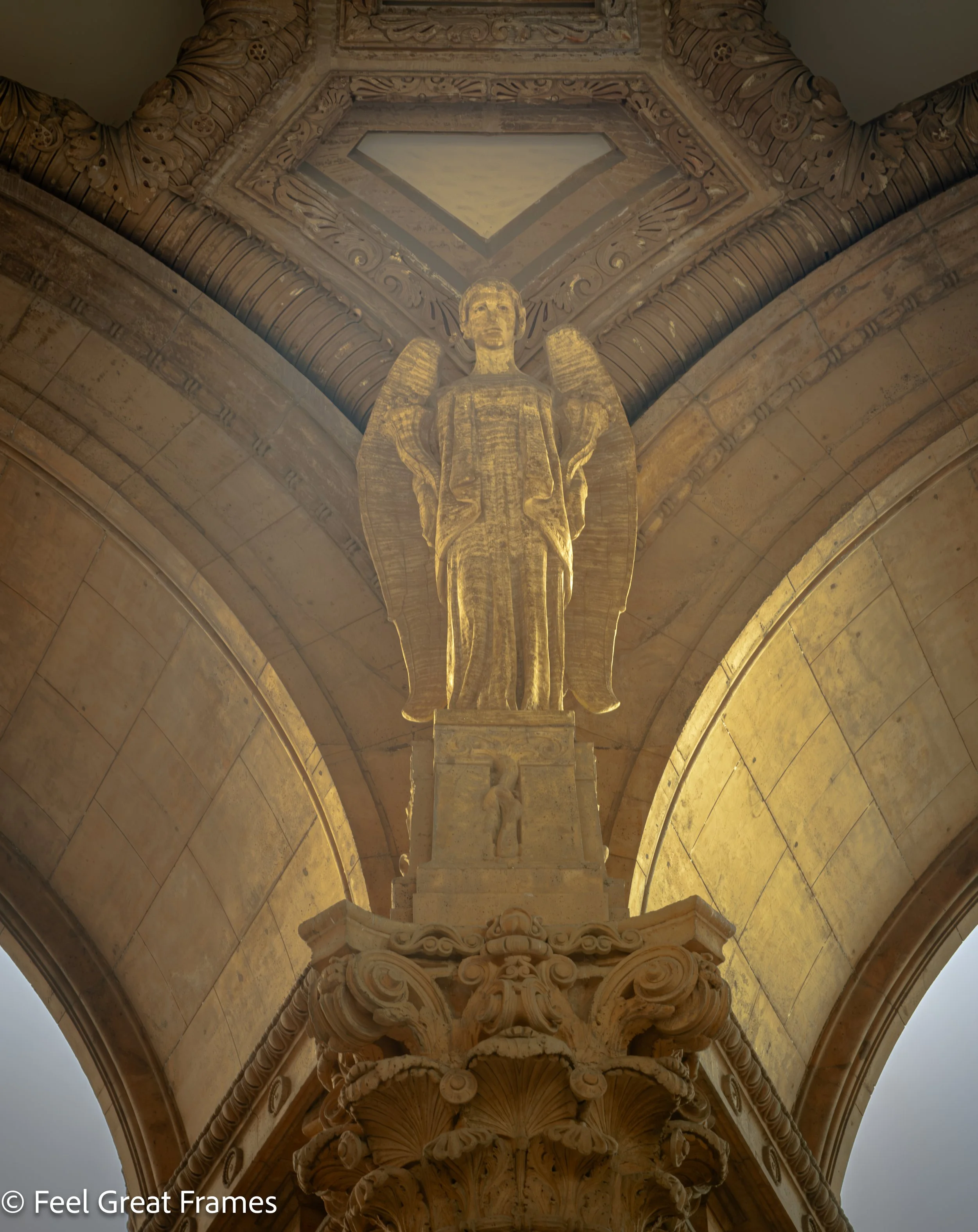 A detailed stone carving of a winged angel figure holding a cross, part of an ornate architectural column inside a church or cathedral, with decorative floral and scroll motifs.