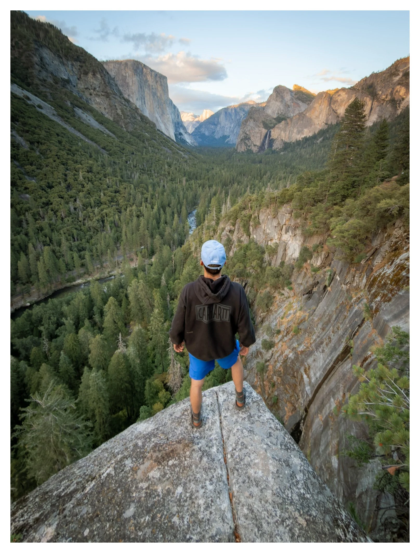 Person standing on a narrow rocky ledge overlooking a lush green valley with waterfall and mountainous cliffs in the background.