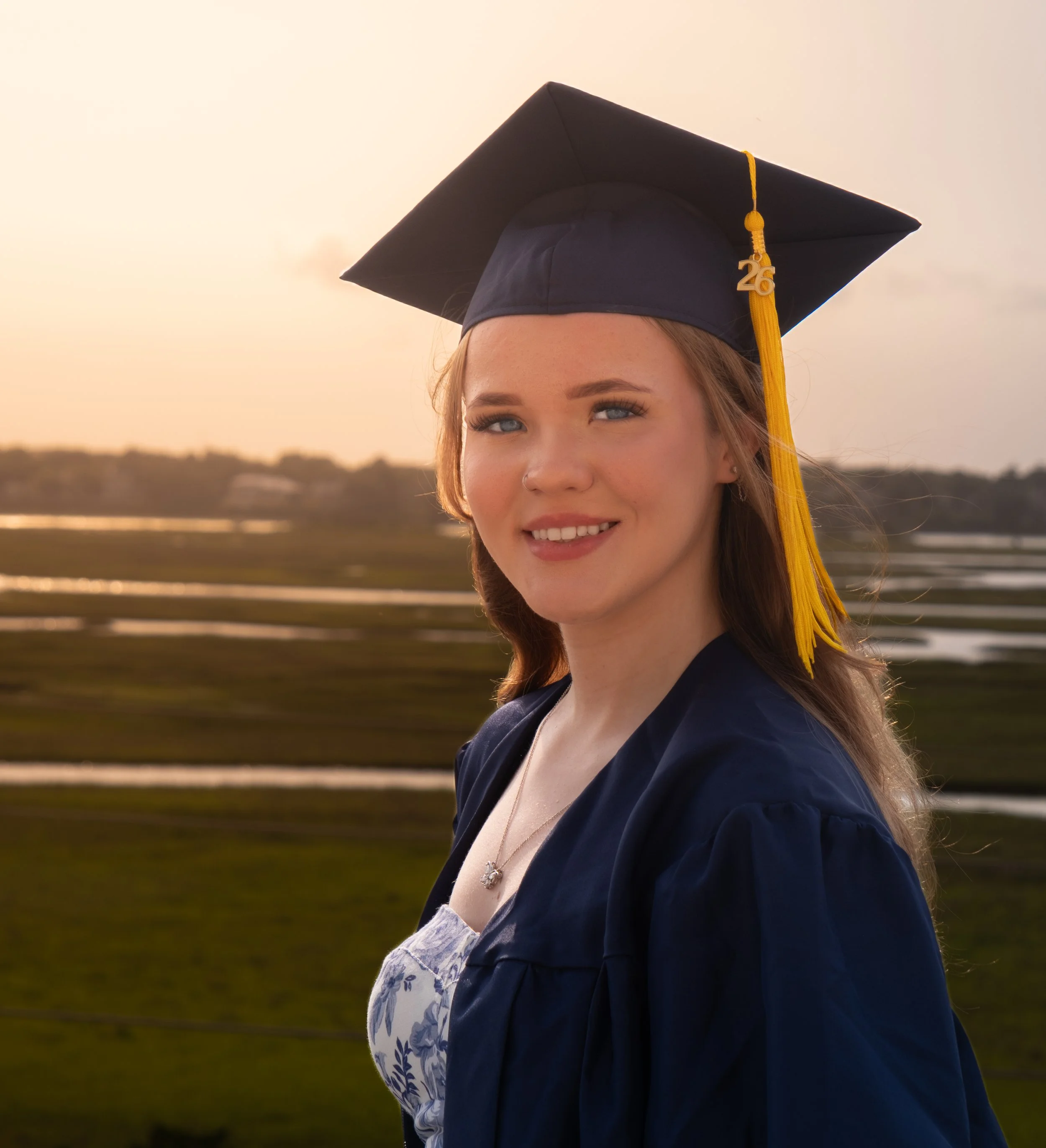 A young woman with blue eyes and fair skin wearing a black graduation cap with a yellow tassel and the year '26' charm, and a navy blue gown, outdoors during sunset with a blurred landscape in the background.
