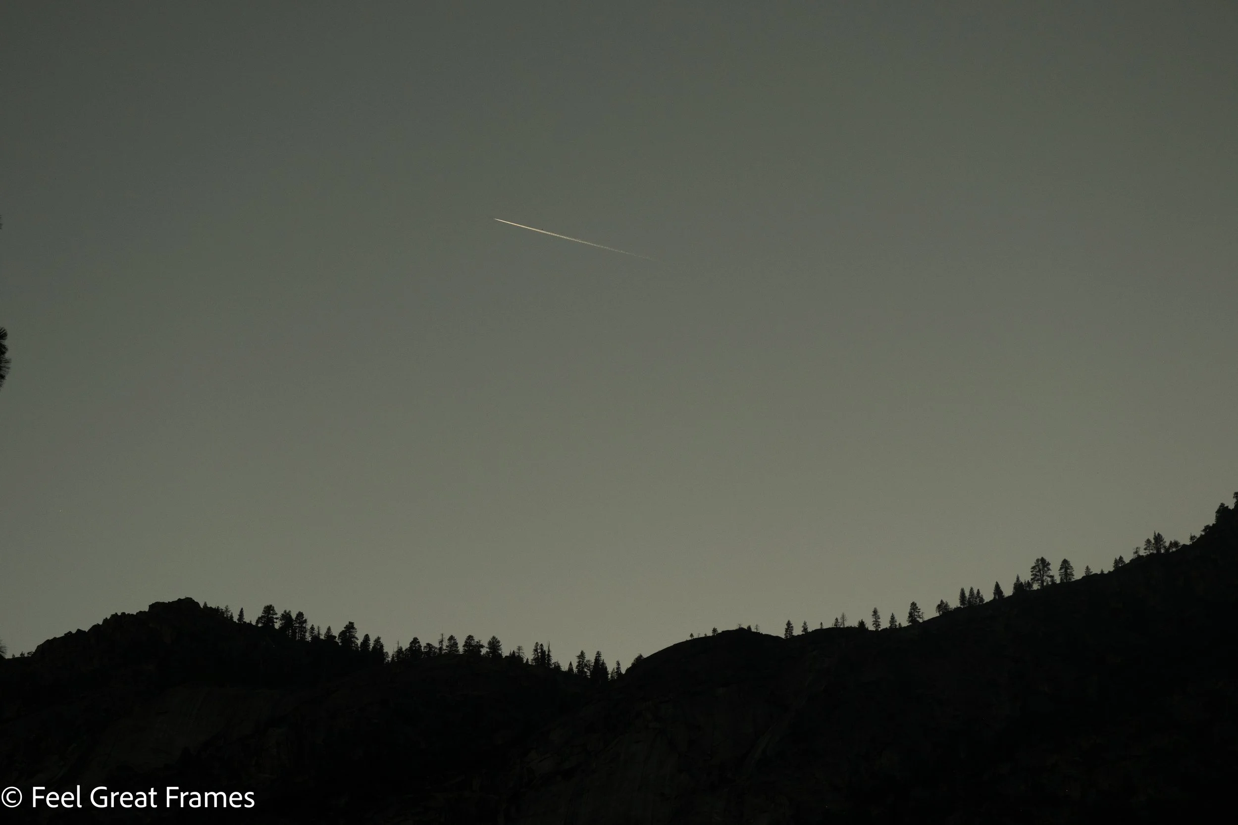 Nighttime mountain landscape with silhouetted hills, trees, and a sky with a faint contrail from an airplane.