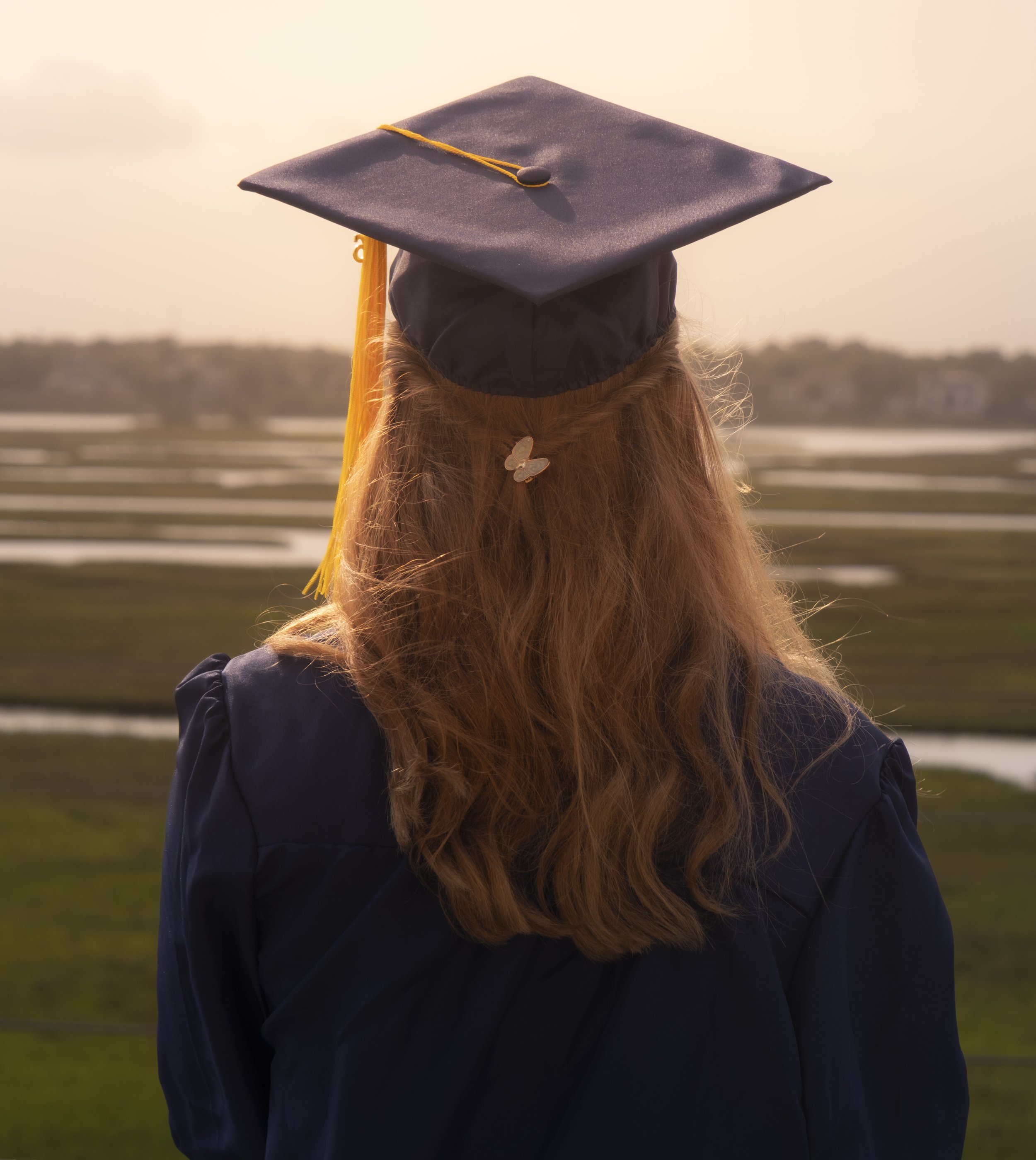 Back view of a person with long red hair wearing a graduation cap and gown, standing outdoors near a body of water with a distant shoreline and sky in the background.