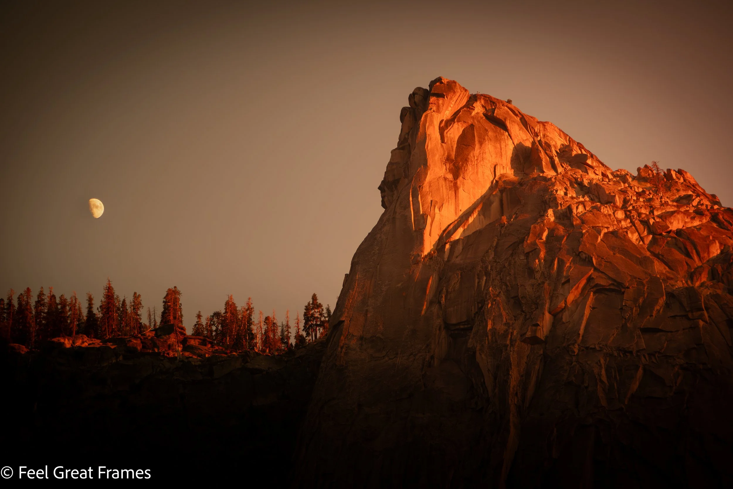 Night view of a mountain illuminated by sunset with the moon in the sky and a forest at the base.