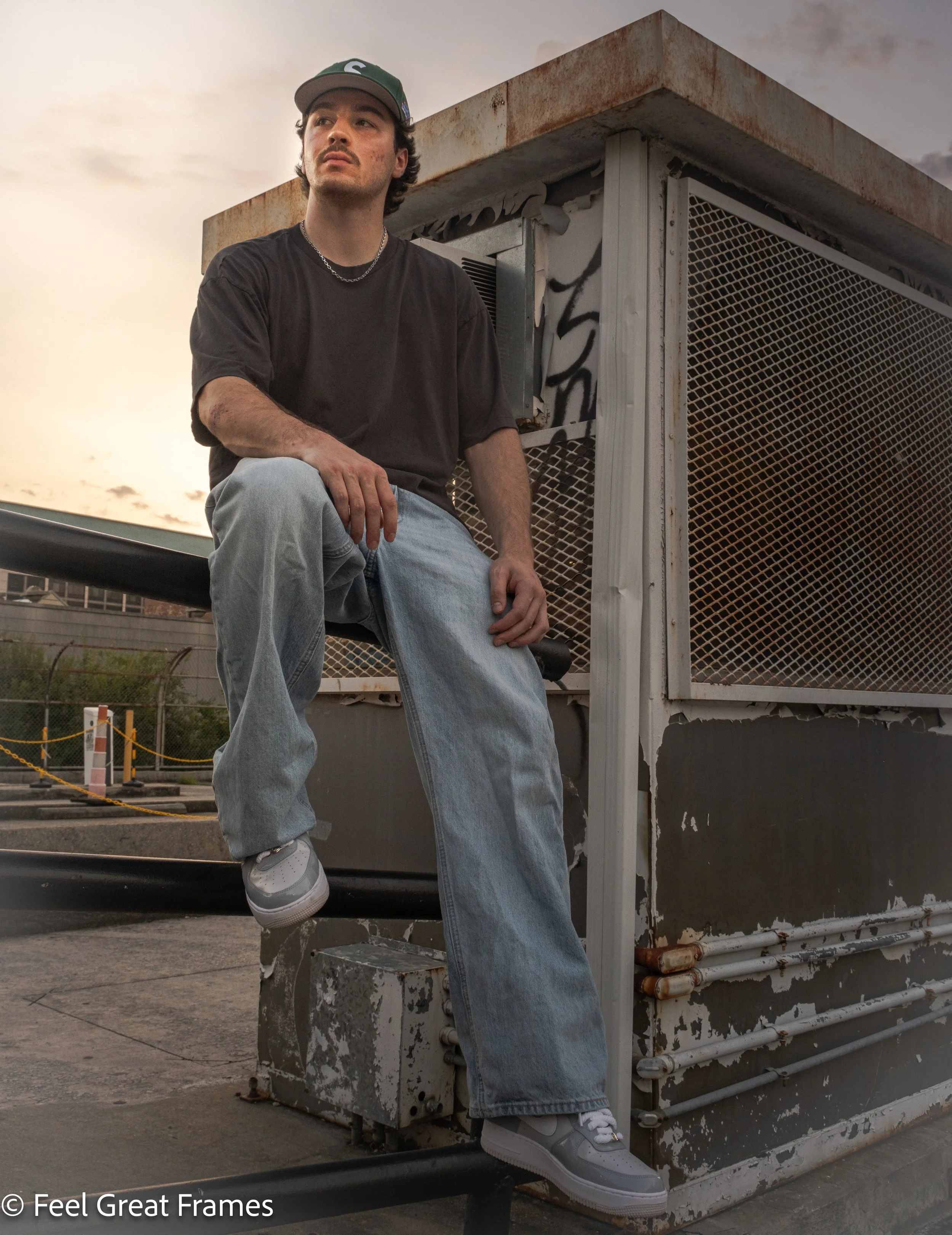 A young man with dark hair, wearing a dark T-shirt, ripped jeans, a chain necklace, gray sneakers, and a dark green cap, sitting on a ledge outdoors at sunset, in front of a rusty industrial structure with graffiti.
