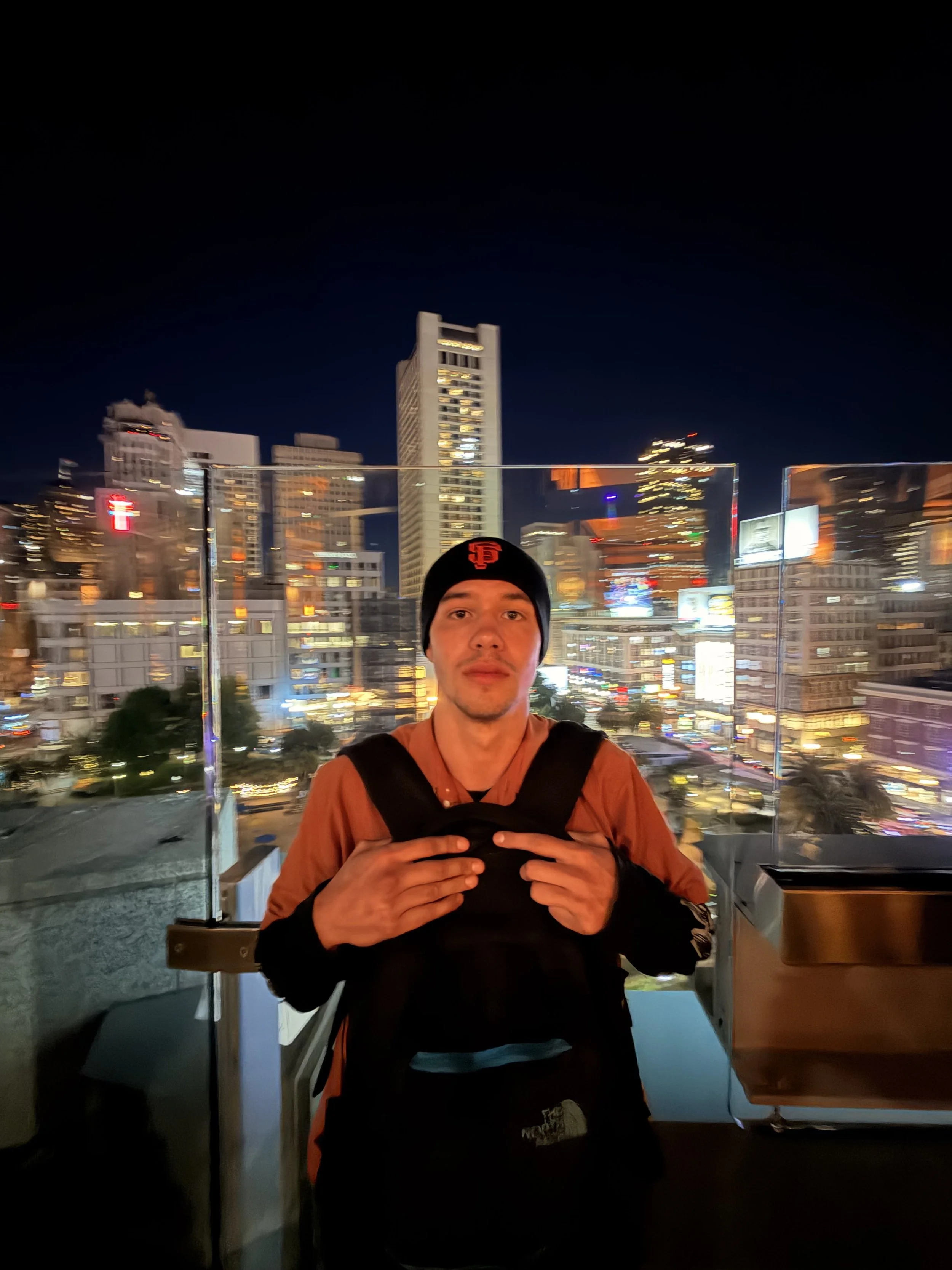 A man wearing a black beanie and orange jacket standing with hands on chest on a rooftop at night, city skyline with illuminated buildings in the background.