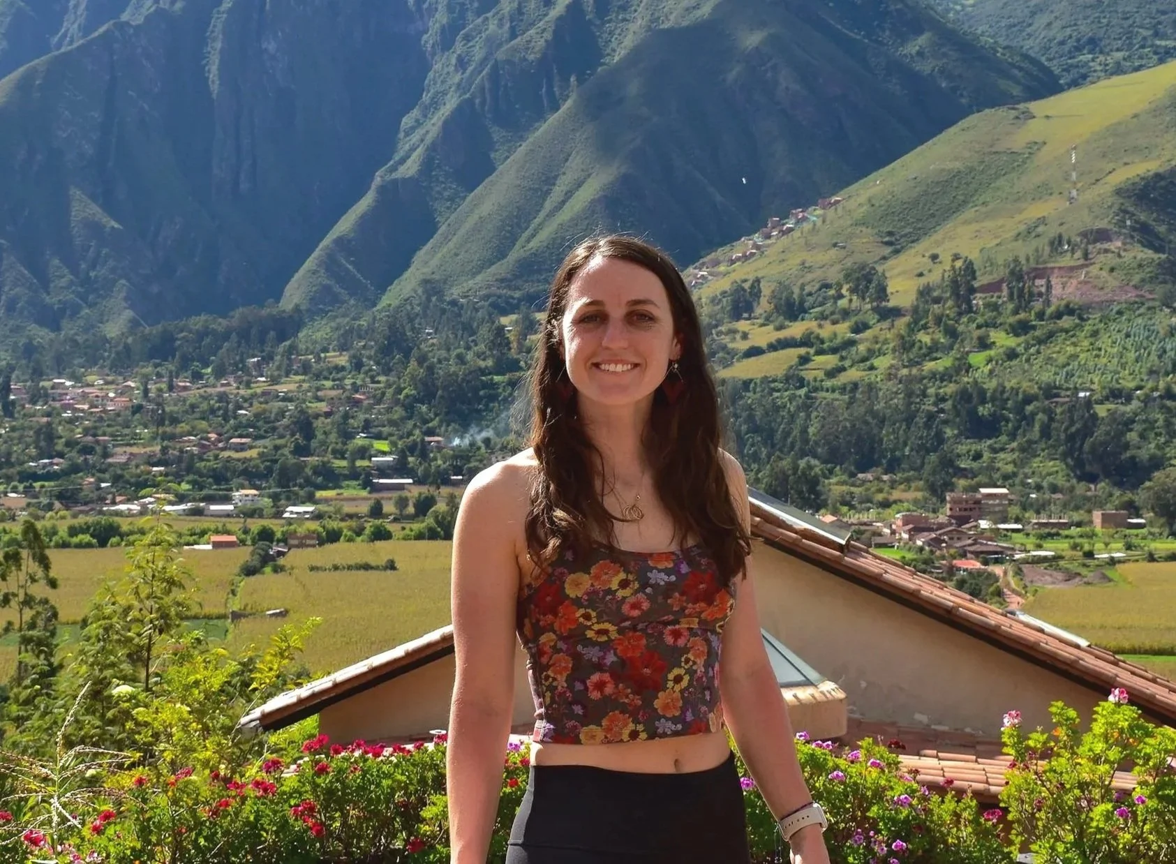 A woman with long brown hair wearing a floral crop top and dark pants, standing on a balcony with mountains and a village in the background, surrounded by colorful flowers.