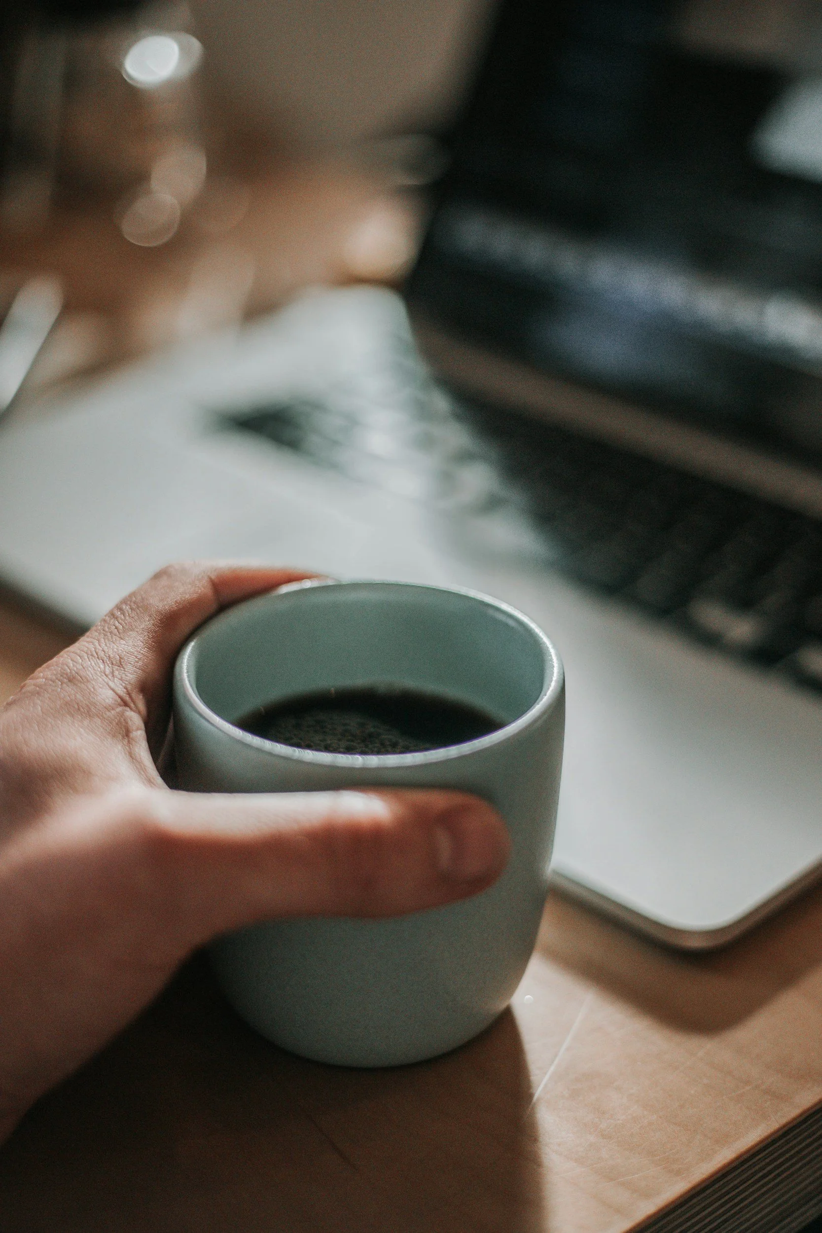 Person holding a grey coffee mug filled with black coffee, viewing a yoga blog on a laptop on a wooden surface in the background.