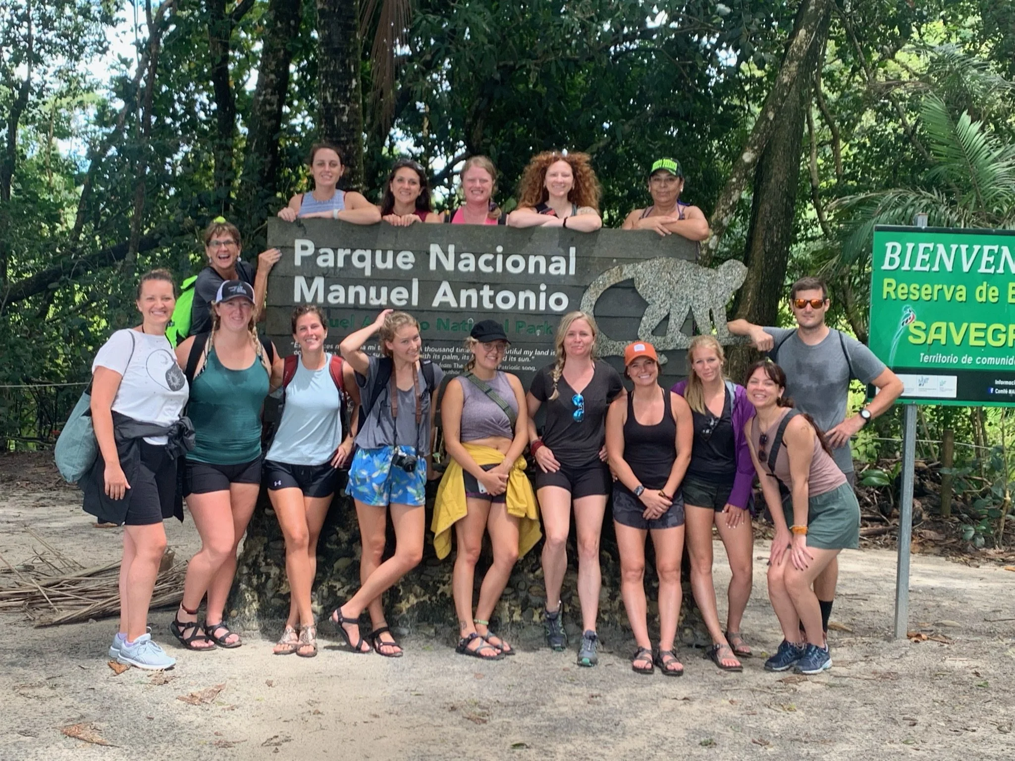 A group of people, mostly women, standing and sitting in front of a sign that reads 'Parque Nacional Manuel Antonio'. They are outdoors with dense jungle trees in the background, and all are dressed in casual summer clothing and footwear, smiling at the camera.
