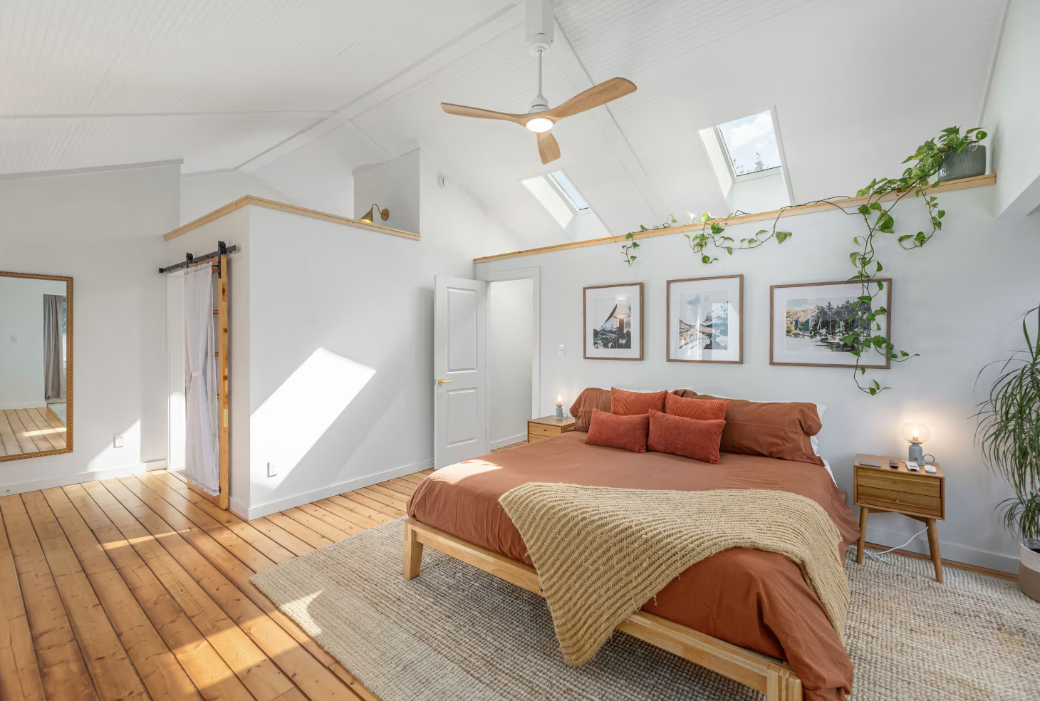 A bright bedroom with white walls, skylights, and a wooden floor. A bed with rust-colored bedding and pillows is centered, with two wooden nightstands on each side. Green plants and framed artwork decorate the wall behind the bed. A ceiling fan and a large mirror are also visible.