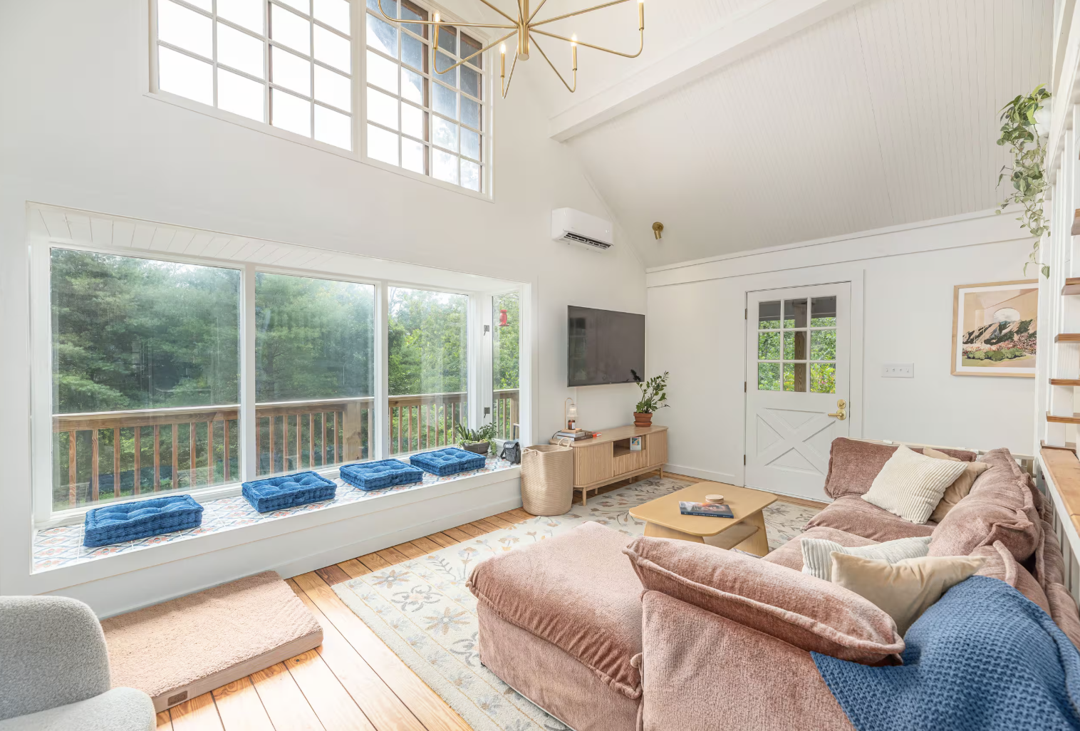 Bright living room with large window seat decorated with blue cushions, white walls, a pink sectional sofa with beige and blue pillows, a wooden coffee table, and a view of greenery outside.