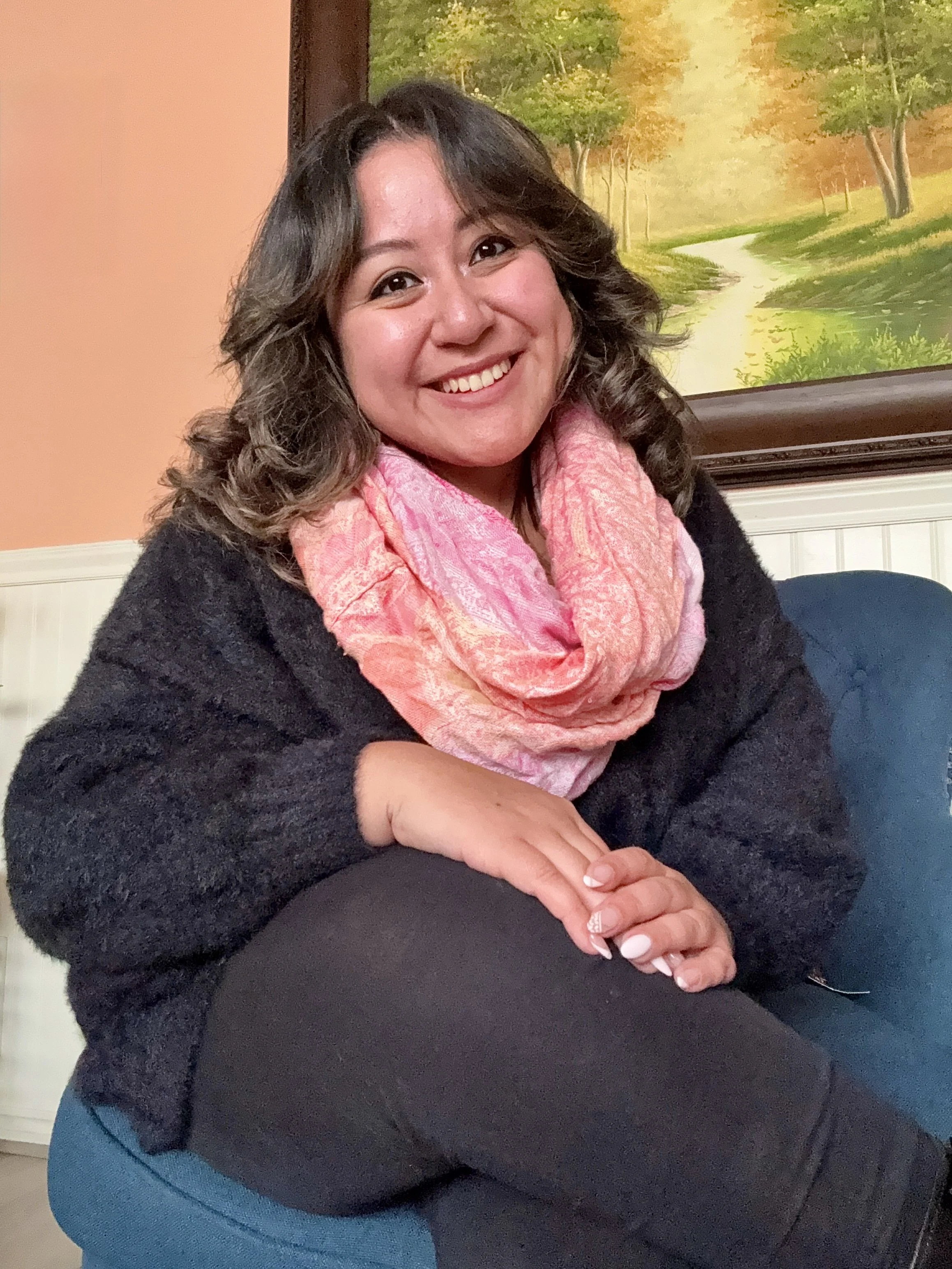 A woman with shoulder-length brown hair, smiling, sitting with her hands folded on her lap. She is wearing a pink scarf, a black sweater, and black pants. There is a framed landscape painting of a pathway through a forest hanging on the wall behind her, and part of a couch or chair is visible.