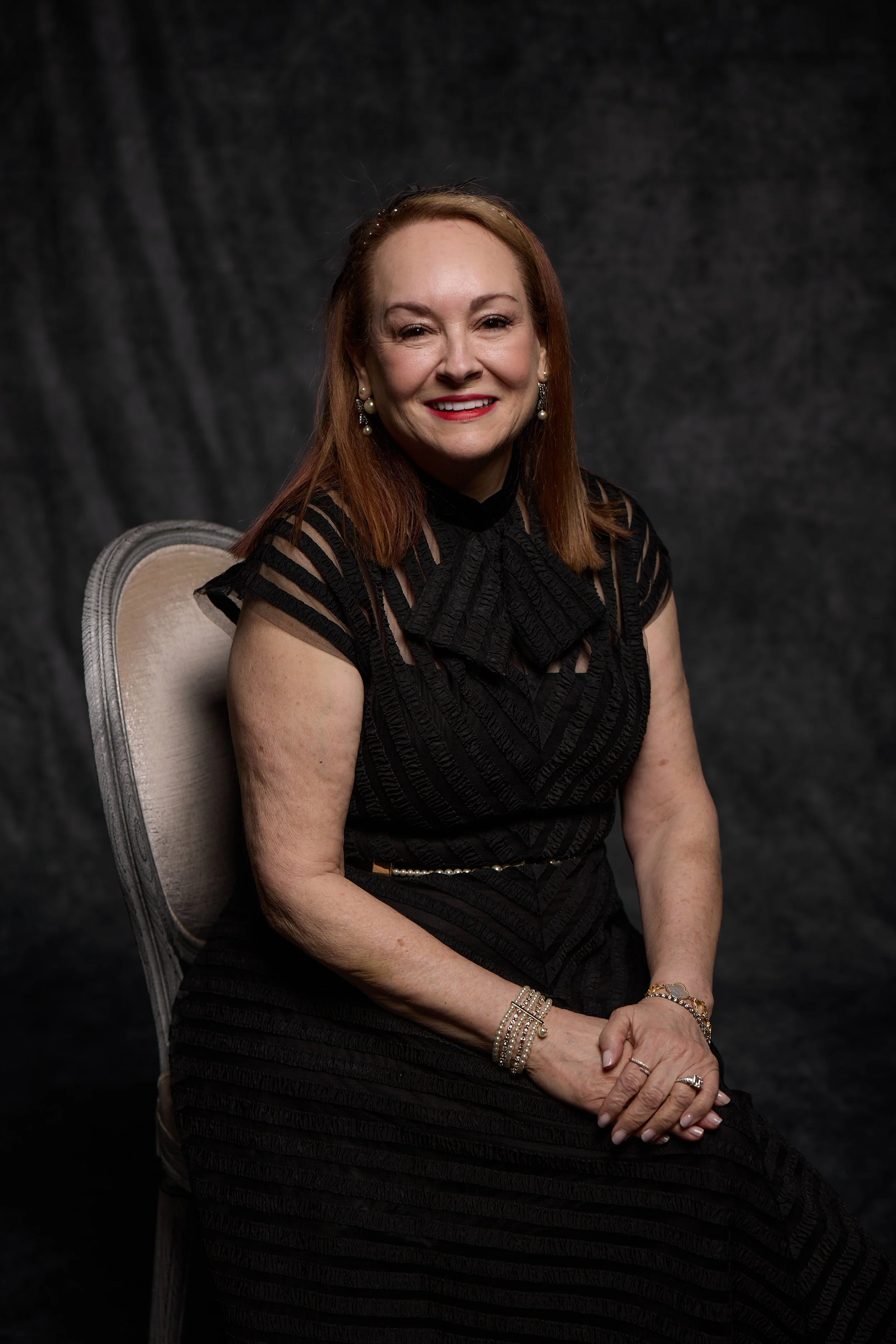 A woman with reddish-brown hair, wearing a black dress with sheer striped patterns, sitting on a beige chair with a rounded back. She is smiling, wearing pearl earrings, a pearl bracelet, a ring, and a gold bracelet. The background is dark and textured.