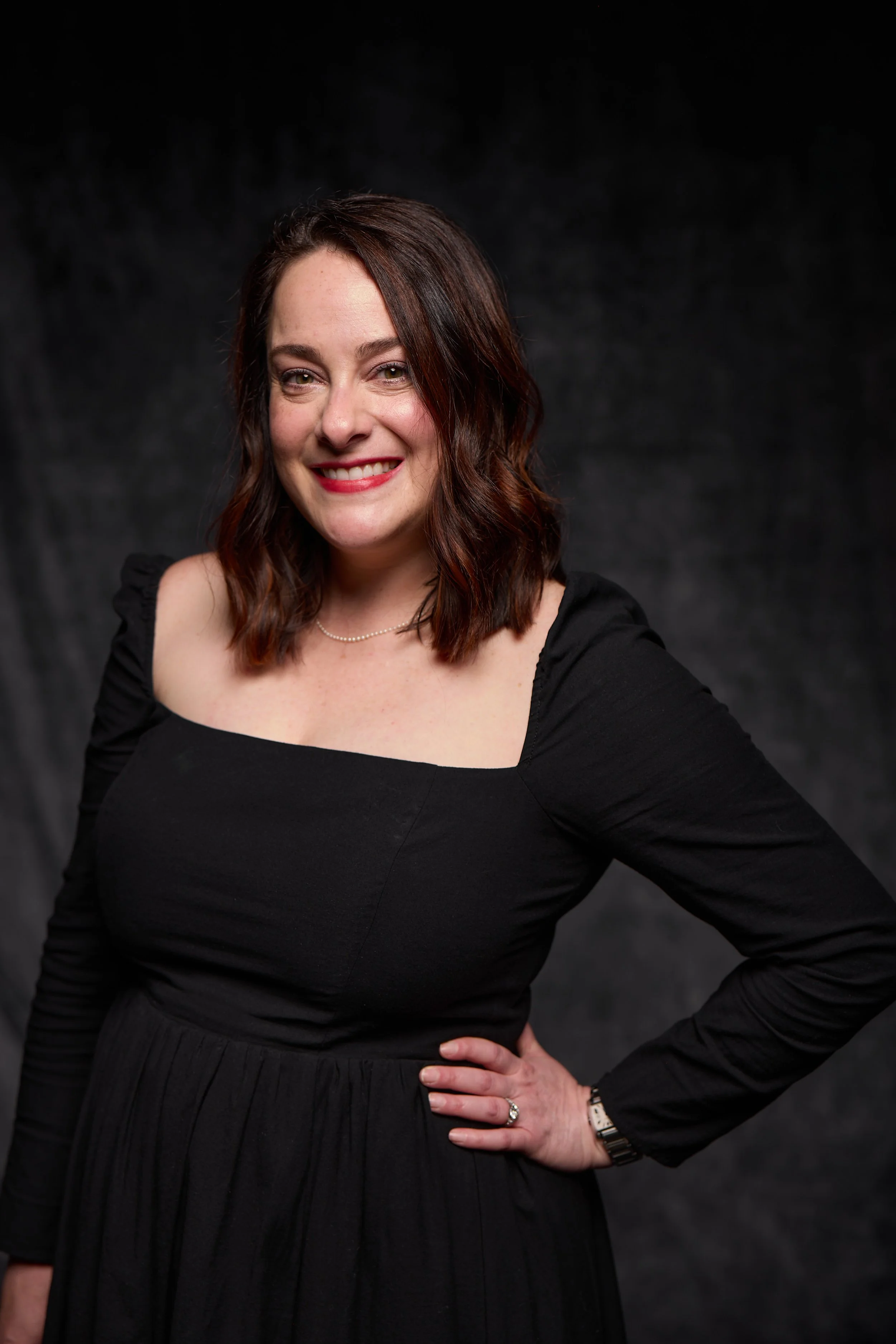 A woman with shoulder-length wavy brown hair smiling and standing against a dark backdrop. She is wearing a black dress with puffed sleeves, a pearl necklace, a watch, and rings. She has one hand on her hip.