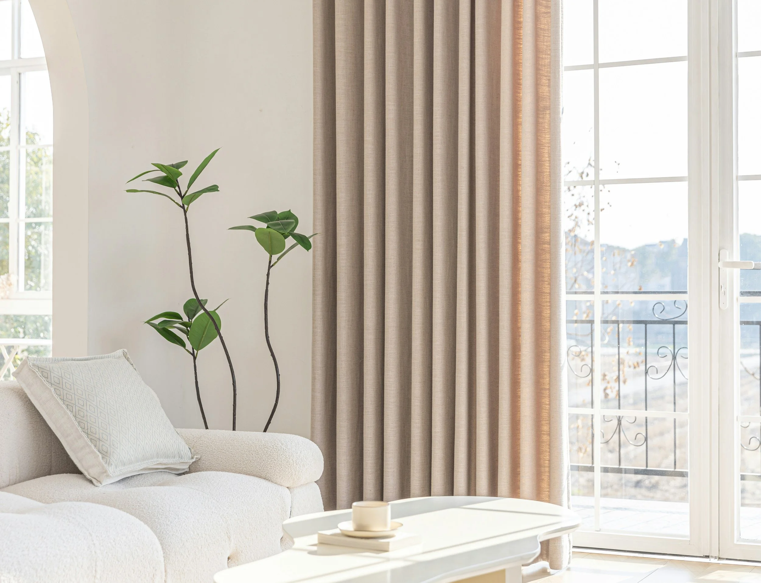 Sunlit living room with white sofa, beige cushions, window with beige curtains, potted plant, and a coffee table with cream-colored candles.