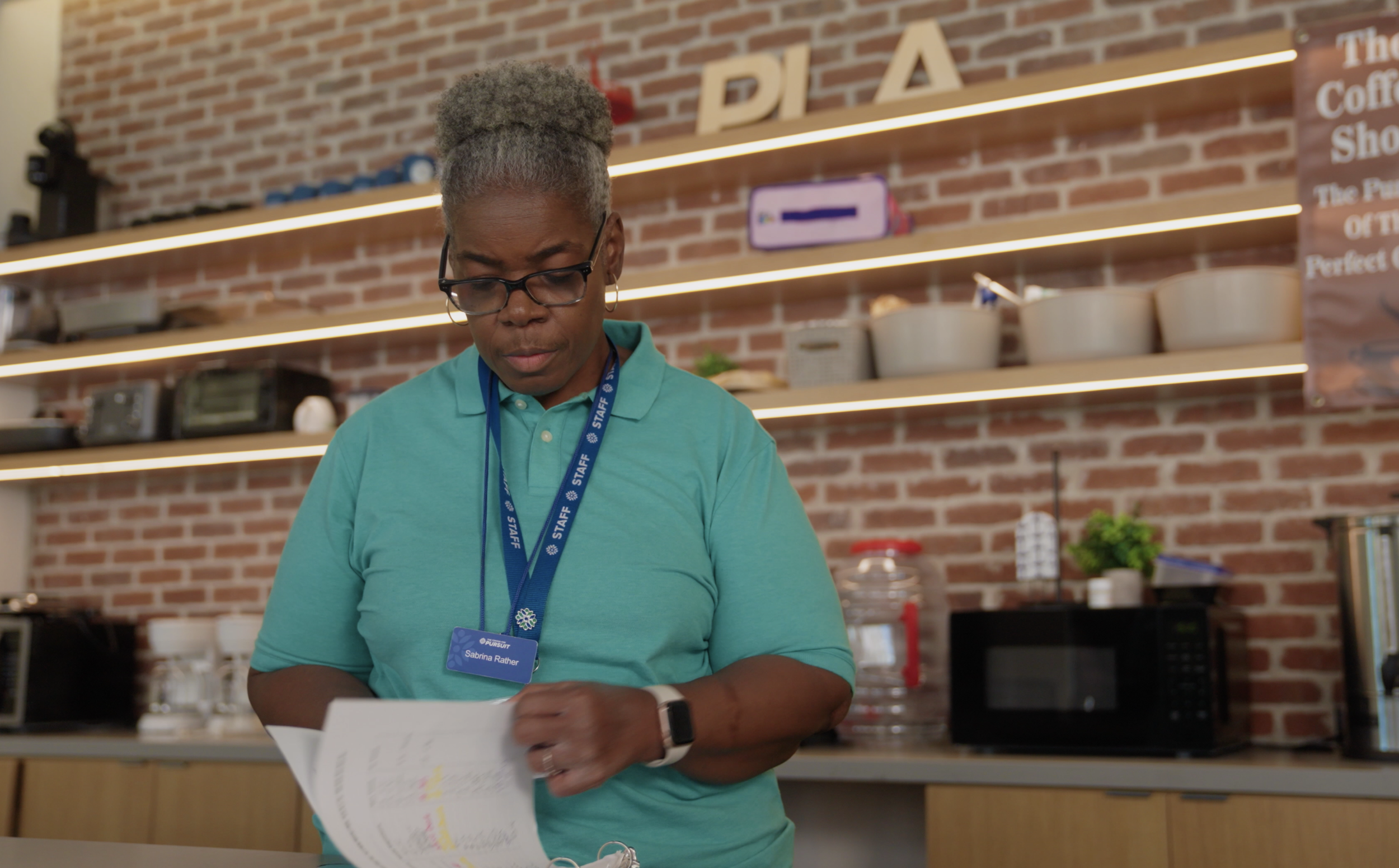 A woman with gray hair styled in an updo, wearing glasses, a teal polo shirt, and a blue staff badge, standing in a coffee shop with exposed brick walls and shelves holding various items.