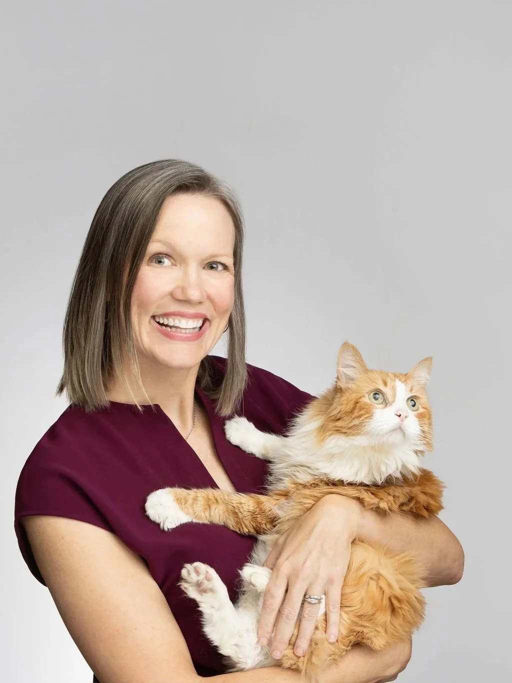 A smiling woman with shoulder-length brown hair wearing a maroon top, holding an orange and white long-haired cat against a plain gray background.