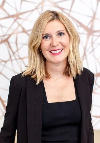 A woman with blonde hair and blue eyes smiling, wearing a black blazer and top, posed in front of a neutral background with wooden geometric design.