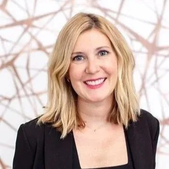 A smiling woman with blonde hair wearing a black blazer against a light background with abstract branch-like patterns.