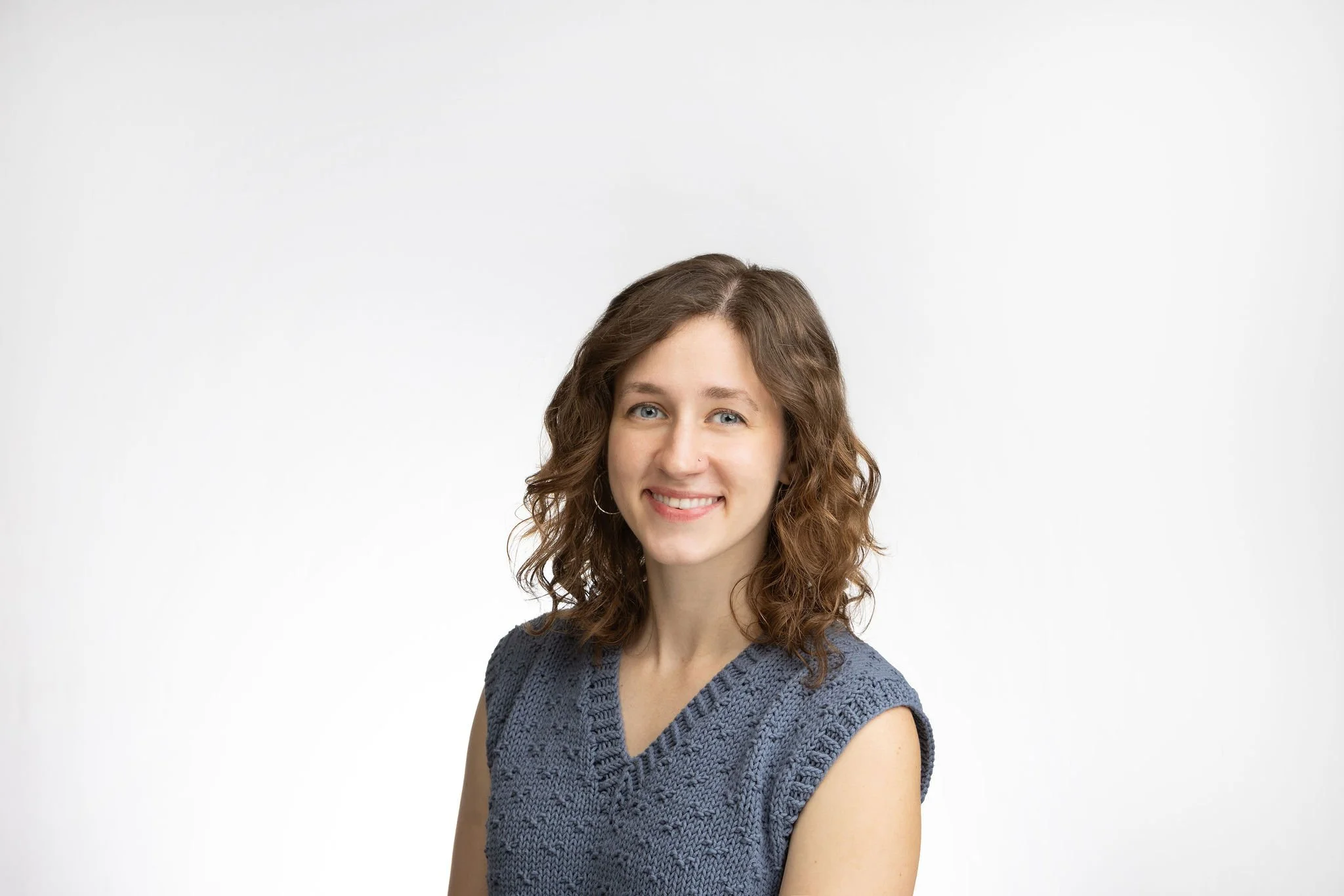 A young woman with wavy brown hair and blue eyes smiling, wearing a knitted gray sleeveless top, standing against a plain white background.